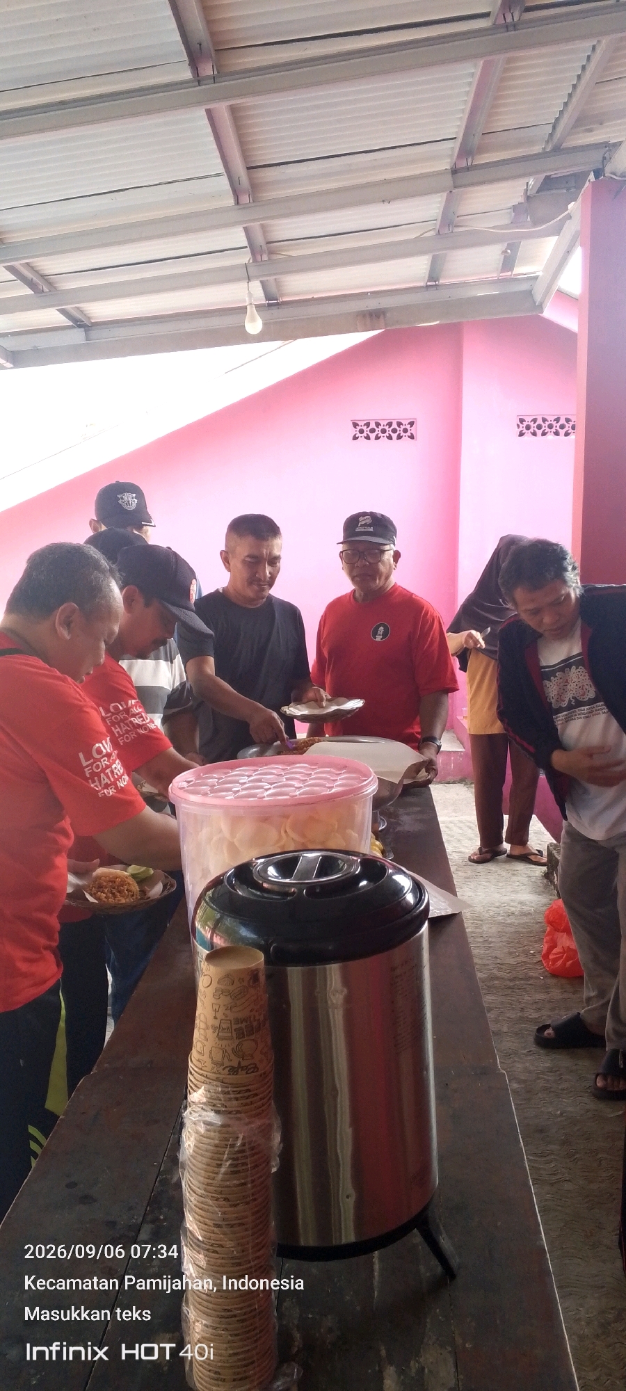 A group of five adult men and one woman are gathered around a buffet-style food service in Pamijahan, Indonesia. The men, dressed in casual attire, are either serving themselves or waiting to be served food from containers on a long table. One man in a red shirt is holding a plate with food, and another man in a black t-shirt is serving food onto a plate. A large pink plastic container filled with crackers is visible, as well as a large stainless steel thermos. In the background, a pink wall with decorative elements and a white corrugated roof are visible. The overall scene suggests an informal gathering or event where food is being distributed. The timestamp indicates the image was taken on 2026/09/06 at 07:34 in Kecamatan Pamijahan, Indonesia.