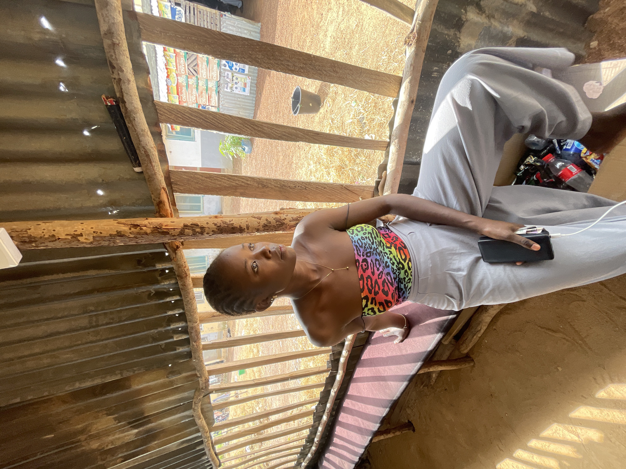 A young woman sits on a wooden bench in a rustic, open-air structure, likely a market stall or a simple shelter. The structure is made of corrugated metal and rough-hewn wooden beams, with sunlight filtering through gaps in the roofing and walls. The woman is wearing a strapless top with a colorful leopard print and light gray wide-leg pants. She holds a smartphone in her right hand, which is plugged into a white charger.
In the background, there are shelves stocked with various items, including what appear to be beverage boxes labeled "Chivita." A dark bucket sits on the ground in the distance. The ground is dusty and dry, with sparse vegetation. The scene suggests a warm, sunny day. The woman's gaze is directed upwards and slightly to her left, with a neutral expression. The overall impression is one of a casual moment in a rural or semi-rural setting in Durimi, Nigeria.