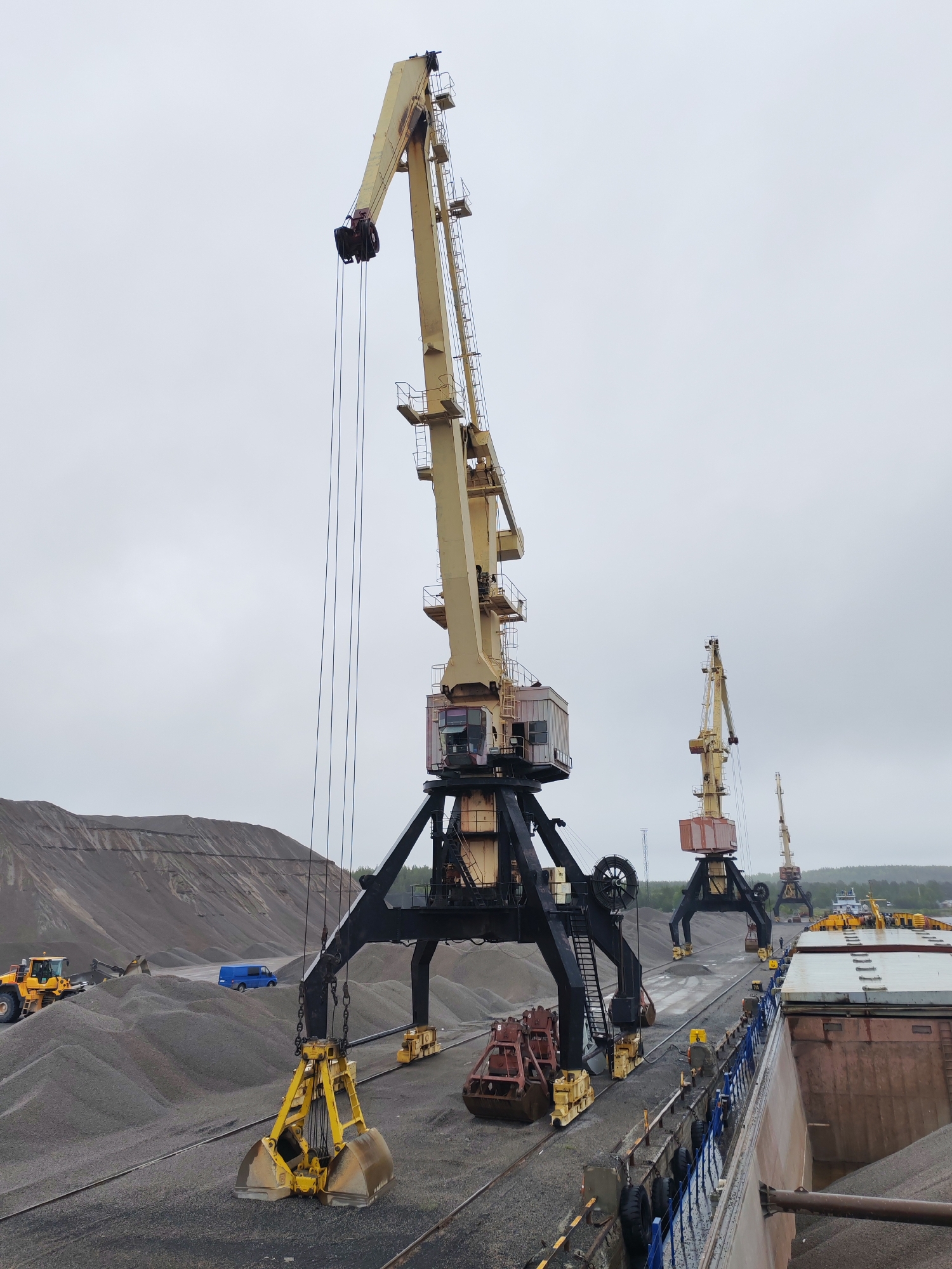 The image depicts a loading dock in Shalsky, Russia, featuring several large, yellow and black port cranes. The cranes are positioned near a pile of gravel or aggregate. A yellow bulldozer is visible near the gravel pile, and a blue van is parked in the background. A ship is docked alongside the loading area, filled with material. The sky is overcast, suggesting a cloudy day.