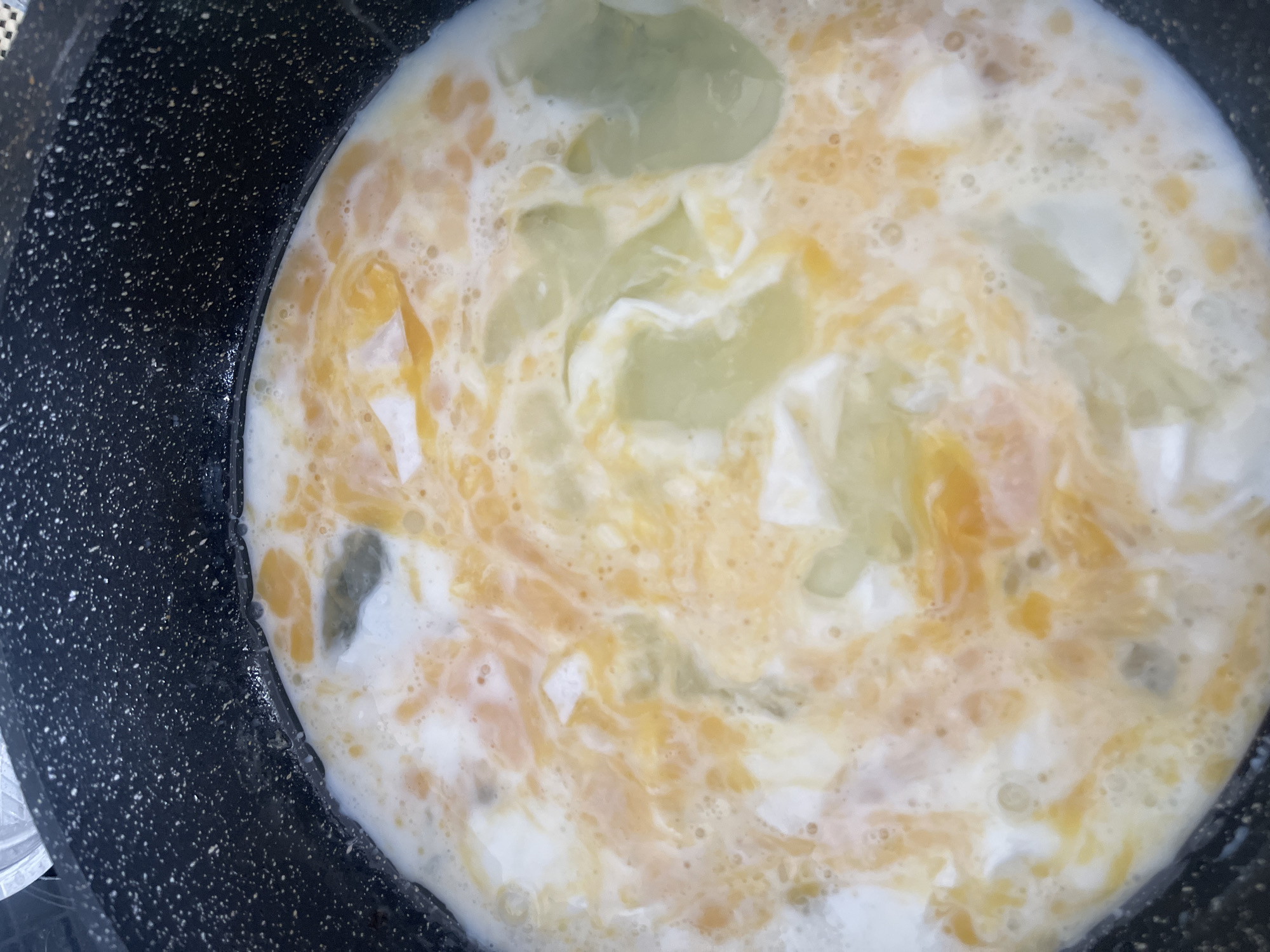This is a close-up, top-down shot of raw eggs being cooked in a black non-stick frying pan. The eggs are partially mixed, with streaks of yellow yolk visible against the white of the egg whites and some larger, translucent, pale green areas which appear to be egg whites that have started to cook. The pan has a speckled texture and a slight sheen, indicating it might be slightly greasy or wet. There are small bubbles forming on the surface of the eggs, suggesting heat is being applied. The scene is indoors, likely a kitchen, but no other environmental details are discernible. There are no people visible in the image. The exact location within Blagoveshchensk, Russia, cannot be determined from the visual information provided.