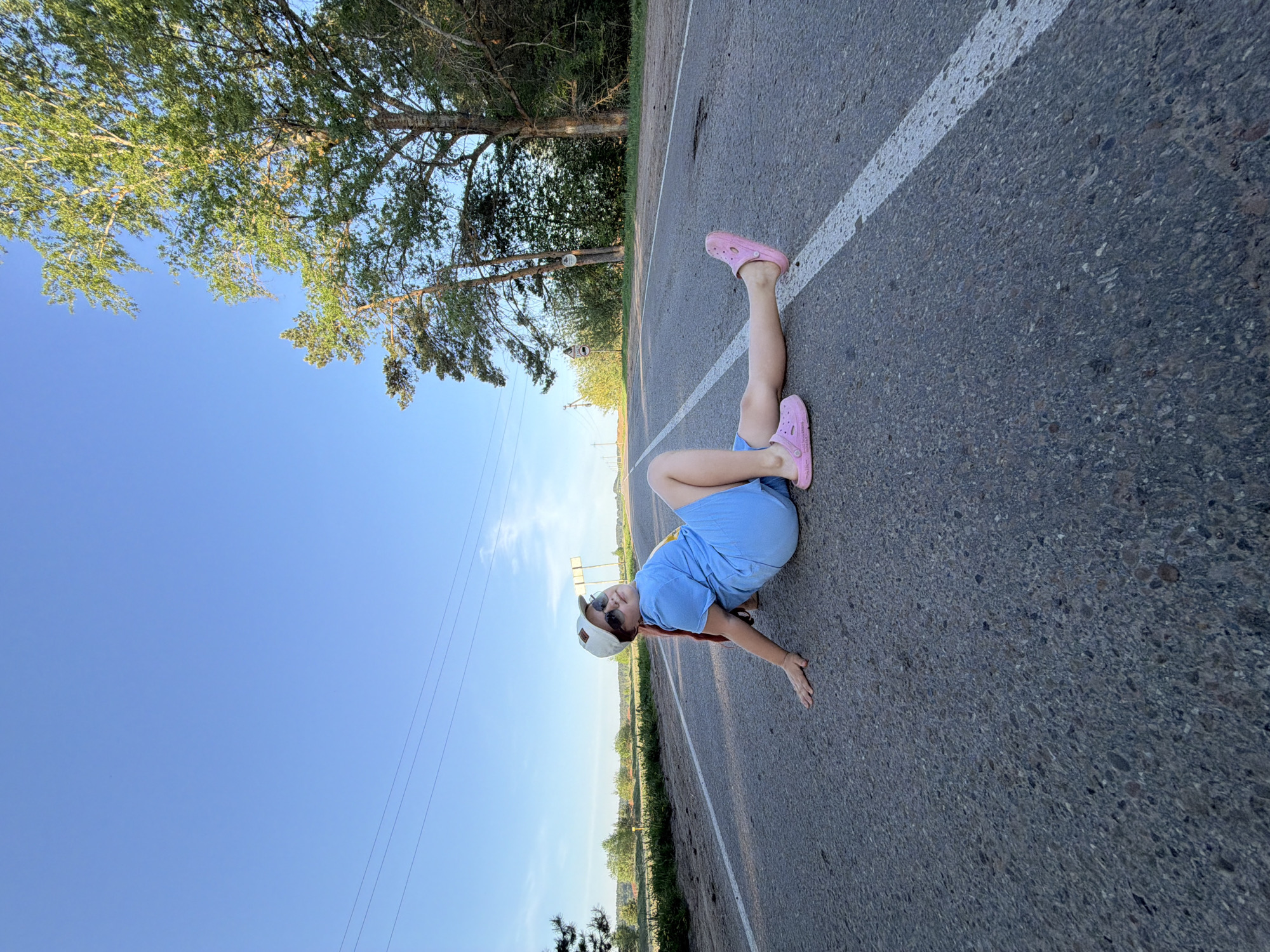 A person is lying on a two-lane road, oriented with their head towards the lower left and their feet towards the upper right of the frame. The individual, dressed in a blue shirt, light blue shorts, and pink Crocs, has a hat and sunglasses on. The road features a single white dividing line.
The sky is blue and clear, while trees line the opposite side of the road. Power lines are visible against the blue sky. The location may be in or near the city of Чумна, Russia, although no specific landmark within the city is evident.