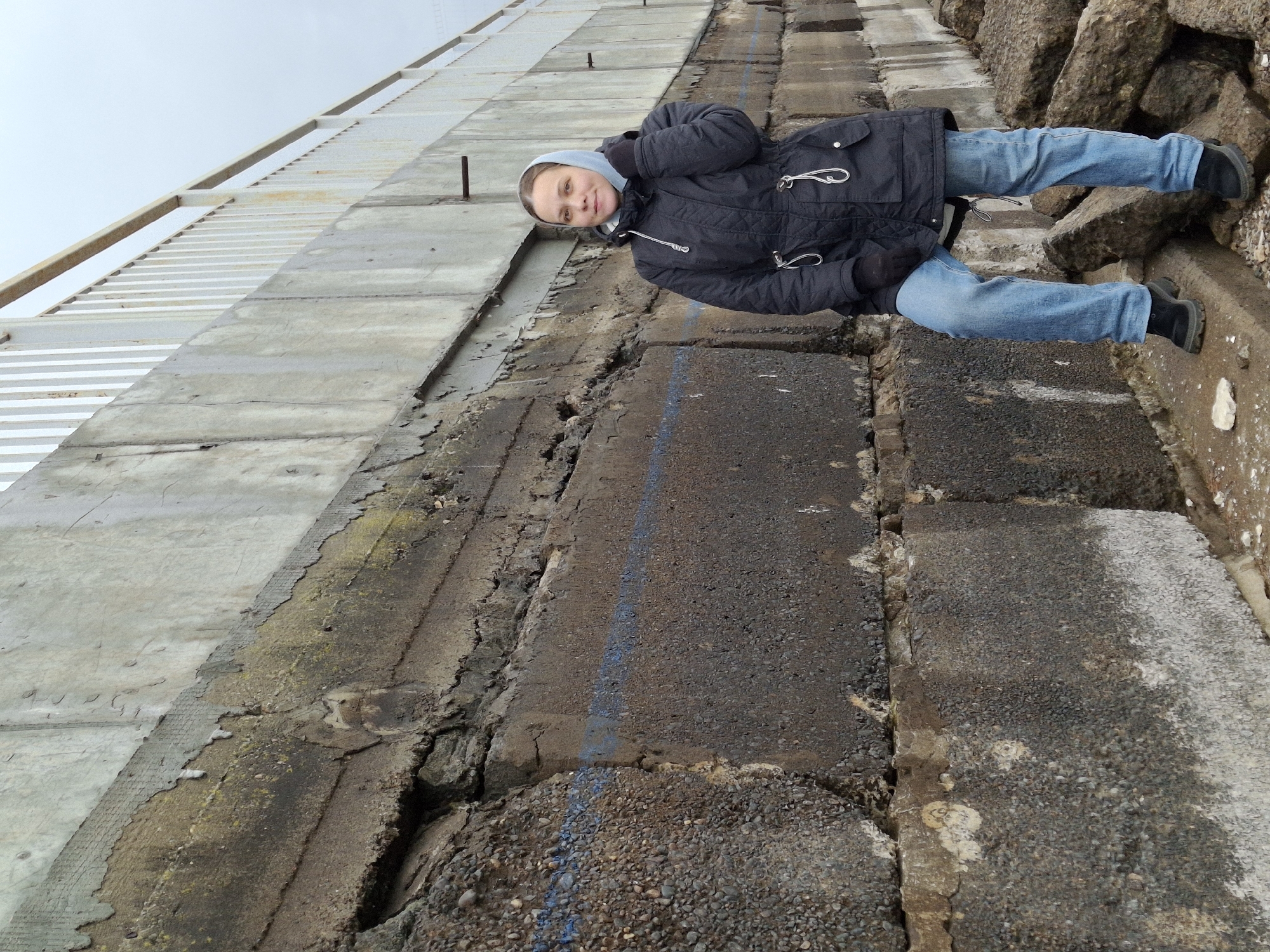 A woman is depicted standing on a rugged, sloping embankment composed of large, dark concrete blocks and rocks. She is wearing a dark blue or black puffer jacket, a light blue hoodie, blue jeans, and dark boots and gloves, indicating cool weather. She looks directly at the camera with a neutral expression, her hands tucked into her jacket.
To her left, a steep, smooth, light grey concrete wall or ramp rises upward. Beyond this wall, a large, light-colored metallic or concrete framework, possibly part of a dam or spillway structure, stretches into an overcast sky. The ground on which she stands is uneven and textured, showing cracked concrete and exposed aggregate. A faint blue painted line runs down the sloped ground in front of her. On the far right, more large, rough-hewn rocks are visible.
The scene suggests an outdoor, possibly industrial or hydro-engineering environment, consistent with areas around a hydroelectric power station or dam, such as the Votkinsk Hydroelectric Power Station in Chaikovsky, Russia. The time of day appears to be daytime, under an overcast sky. A small, faint white marking, possibly "L3," is visible on a light patch on one of the concrete slabs in the lower right foreground. No other visible text or distinct activity is present.