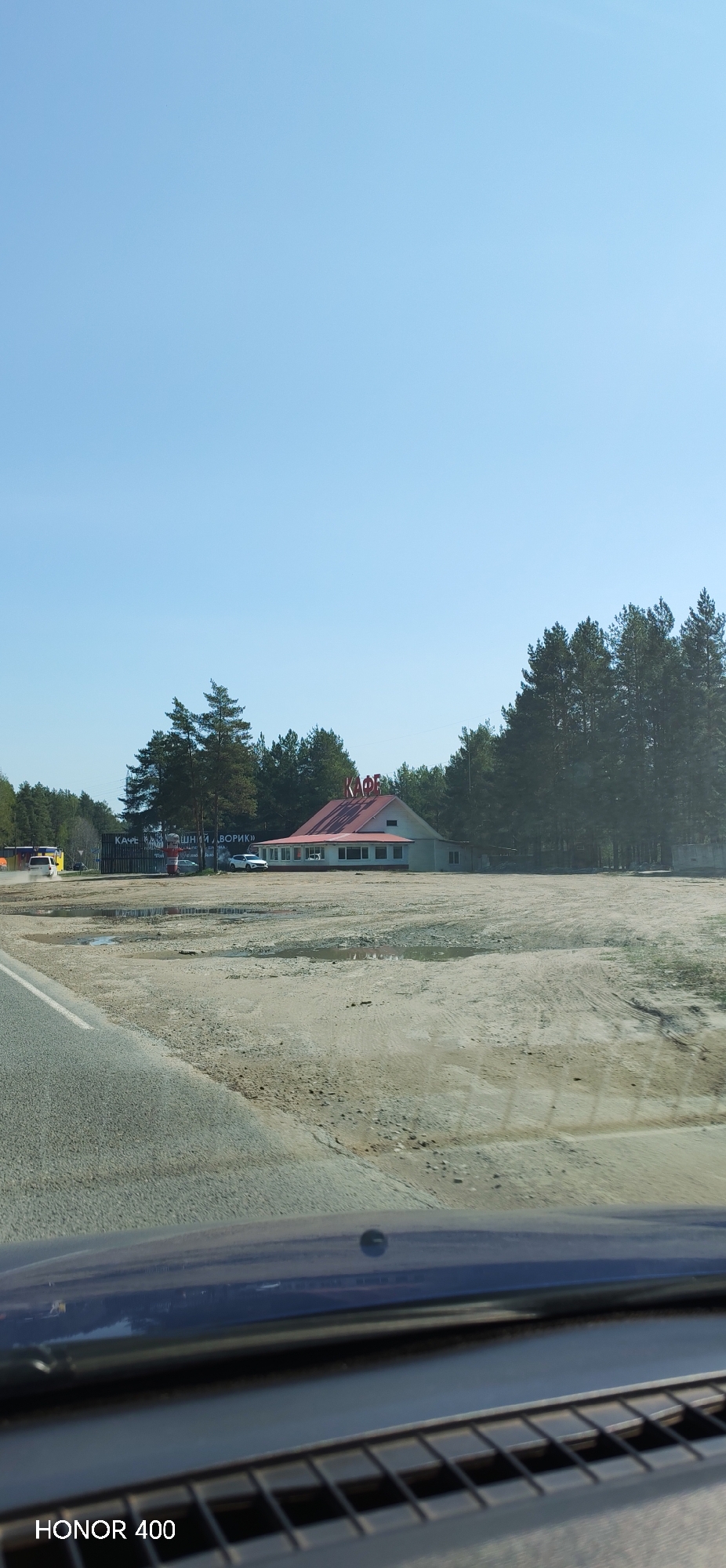 The image depicts a scene from inside a vehicle, possibly a car, looking out towards a cafe situated near a forest. The cafe, identified by the Cyrillic word "КАФЕ" (CAFE) on its roof, has a red roof and white walls. It is located on a dusty, sandy lot with patches of standing water. Behind the cafe, there is a dense forest with tall, mature trees. The sky is clear and blue. In the foreground, the dashboard of the vehicle is visible. A watermark indicating "HONOR 400" is present at the bottom left corner of the image. The location is indicated as Noviy, Russia, likely a rural or semi-rural area given the surrounding landscape. There are cars and structures visible next to the cafe.