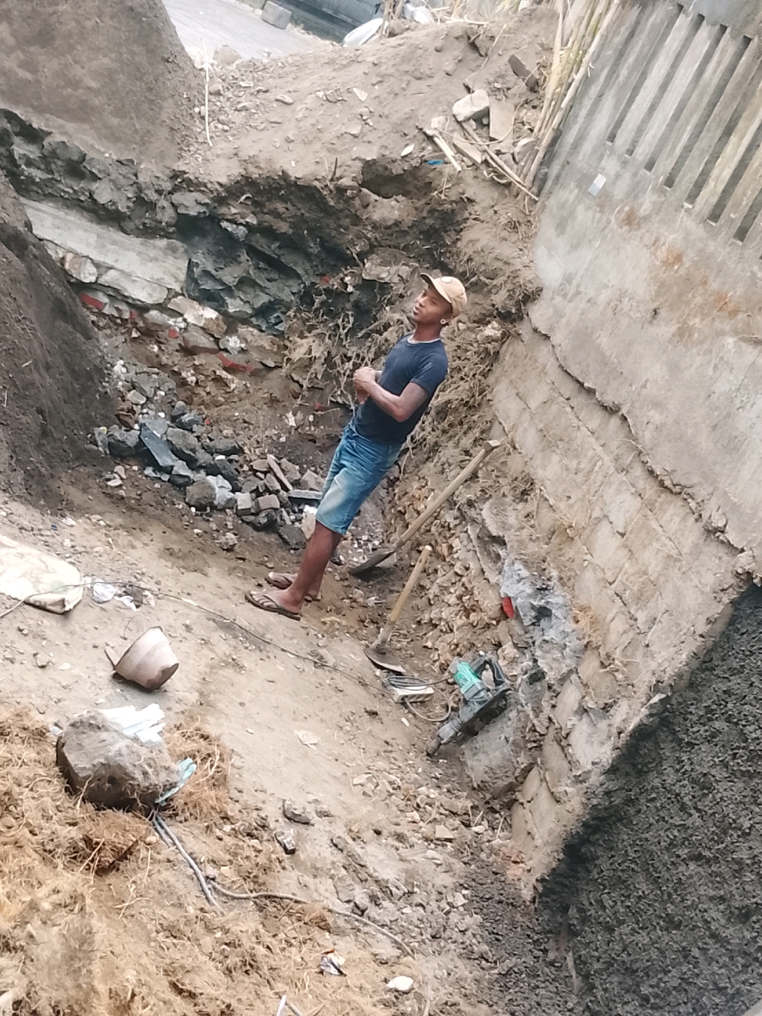 A male construction worker, wearing a dark t-shirt, denim shorts, and a light-colored baseball cap, stands in a dirt excavation site. He is looking upwards with his arms crossed. Nearby, a shovel and a jackhammer are visible on the ground. The site appears to be undergoing construction or demolition, with piles of dirt, rocks, and debris present. The background includes a concrete structure and a road. The location is identified as Kuta, Indonesia.