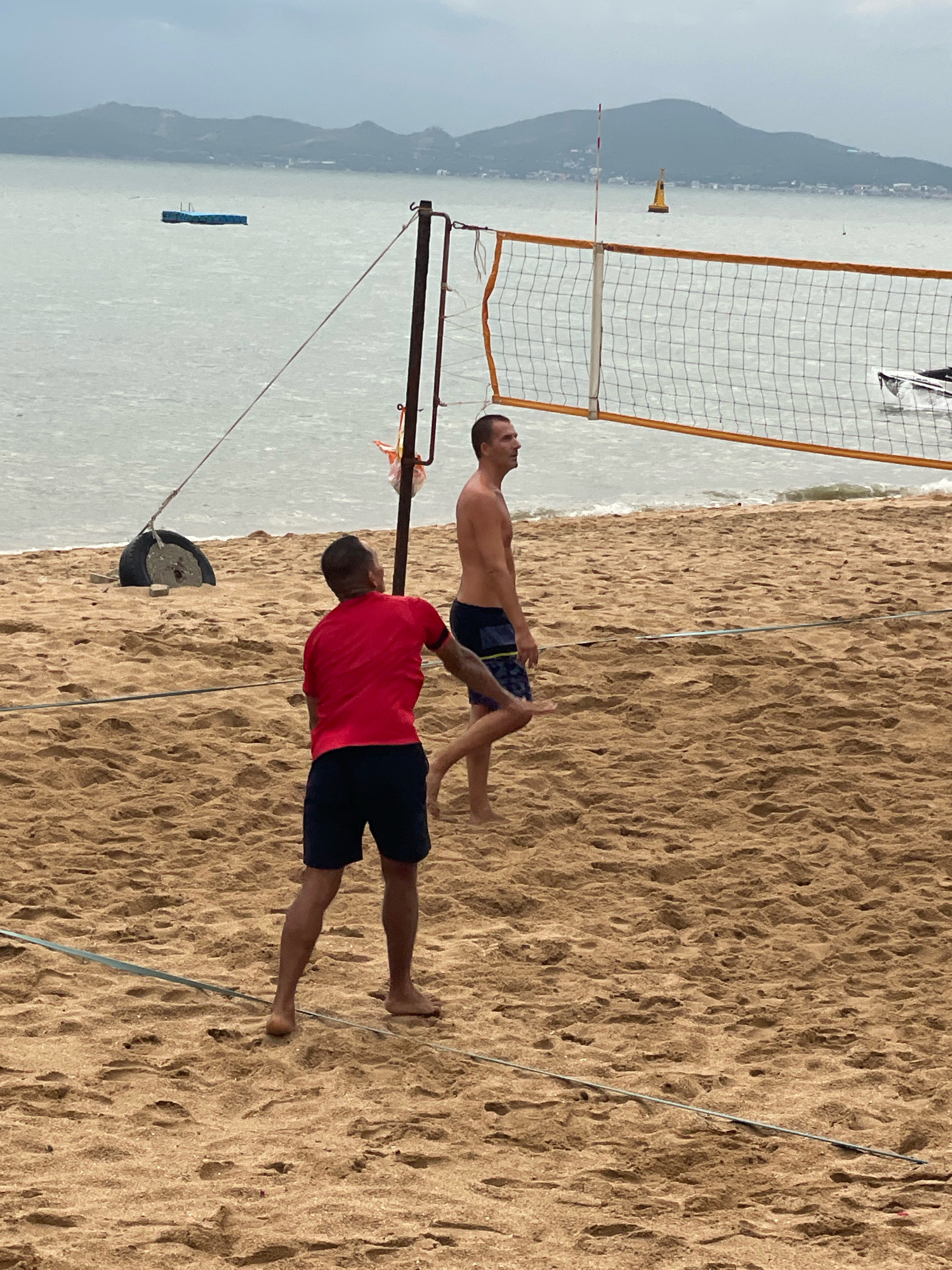 The image depicts a beach volleyball court on a sandy beach. Two shirtless men are playing volleyball. One man, wearing a red t-shirt and dark shorts, is facing away from the camera, appearing to be in the middle of a game. The other man, wearing dark patterned shorts, is facing the net. A volleyball net is set up on the court, with poles and tensioning ropes visible. In the background, the ocean stretches out to a hazy horizon, with distant mountains and a few small boats. The sky is overcast, suggesting a cloudy day. The location is identified as Pattaya City, Thailand.