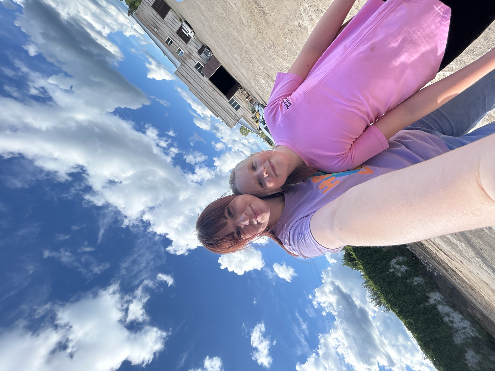 Two females are taking a selfie. The location appears to be Chaikovsky, Russia, based on architectural styles common to the region. The younger female is wearing a pink shirt and the older female is wearing a purple shirt. The background features a blue sky with scattered clouds and a building with metal siding. There is also greenery present, suggesting a rural or suburban environment.
