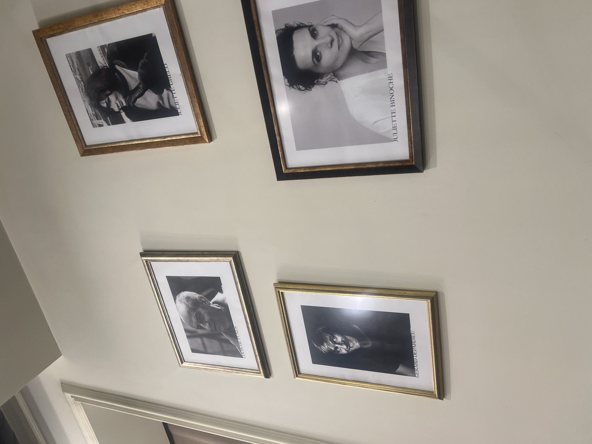 The media file captures an interior scene in Yerevan, Armenia, featuring a light-colored wall adorned with four framed black and white portraits of notable French personalities. The image itself is rotated, making the frames appear diagonally oriented on the wall.
In the upper section, two portraits are displayed. On the left, a gold-framed picture shows a black and white portrait of a woman, with the name "JULIETTE GRECO" printed vertically along the right side of the image within the frame. To its right, a larger black frame with a gold inner trim holds a black and white portrait of Juliette Binoche, with her name "JULIETTE BINOCHE" also printed vertically along the right edge.
In the lower section of the wall, two more portraits are visible. On the left, a silver-gold framed portrait features Louis de Funès, identified by his name "LOUIS DE FUNÈS" printed vertically along the right side. To its right, another gold-framed portrait displays Gérard Depardieu, with his name "GERARD DEPARDIEU" visible along the right edge.
All portraits are black and white headshots or upper-body shots. The wall is plain and light-colored, suggesting a neutral interior setting like a hallway or room. A white architectural molding or door frame is partially visible at the bottom of the image. There is no activity taking place; the image is a static capture of wall decor.