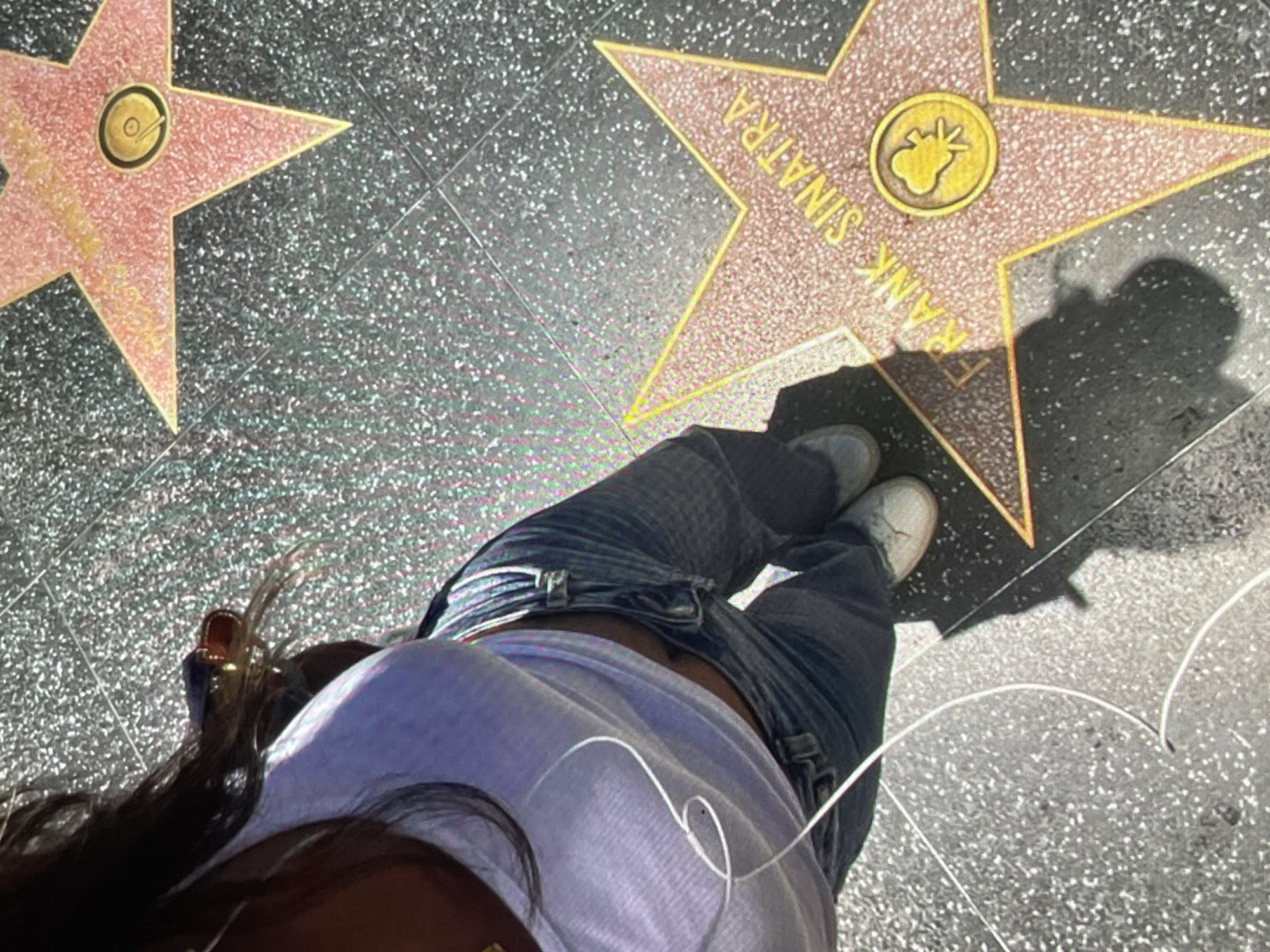The image captures a high-angle, first-person perspective of a person's lower torso and legs, looking down at the Hollywood Walk of Fame. The individual is wearing dark blue jeans, white sneakers, and a light purple, possibly cropped, short-sleeved top. White wired earphones are draped across the person's front. Long dark hair is visible, with a gold-colored hair clip or accessory near the shoulder.
The setting is an outdoor pavement featuring two pink terrazzo stars with brass borders and lettering, characteristic of the Hollywood Walk of Fame. The star prominently positioned on the right side of the frame clearly displays the name "FRANK SINATRA" in gold letters. Below the name, a circular brass emblem features a phonograph symbol, indicating his contribution to the recording industry. A strong, distinct shadow of a person, likely the photographer, is cast across the "FRANK SINATRA" star and the surrounding pavement, indicating bright, sunny daylight. A second, partial star is visible on the upper left side of the frame. The dark gray pavement itself has a glittering, speckled texture.
The activity depicted is simply standing or walking on this famous landmark. Notable details include the clear identification of Frank Sinatra's star, the sunny weather, and the distinctive appearance of the Walk of Fame. The visible text is "FRANK SINATRA."
