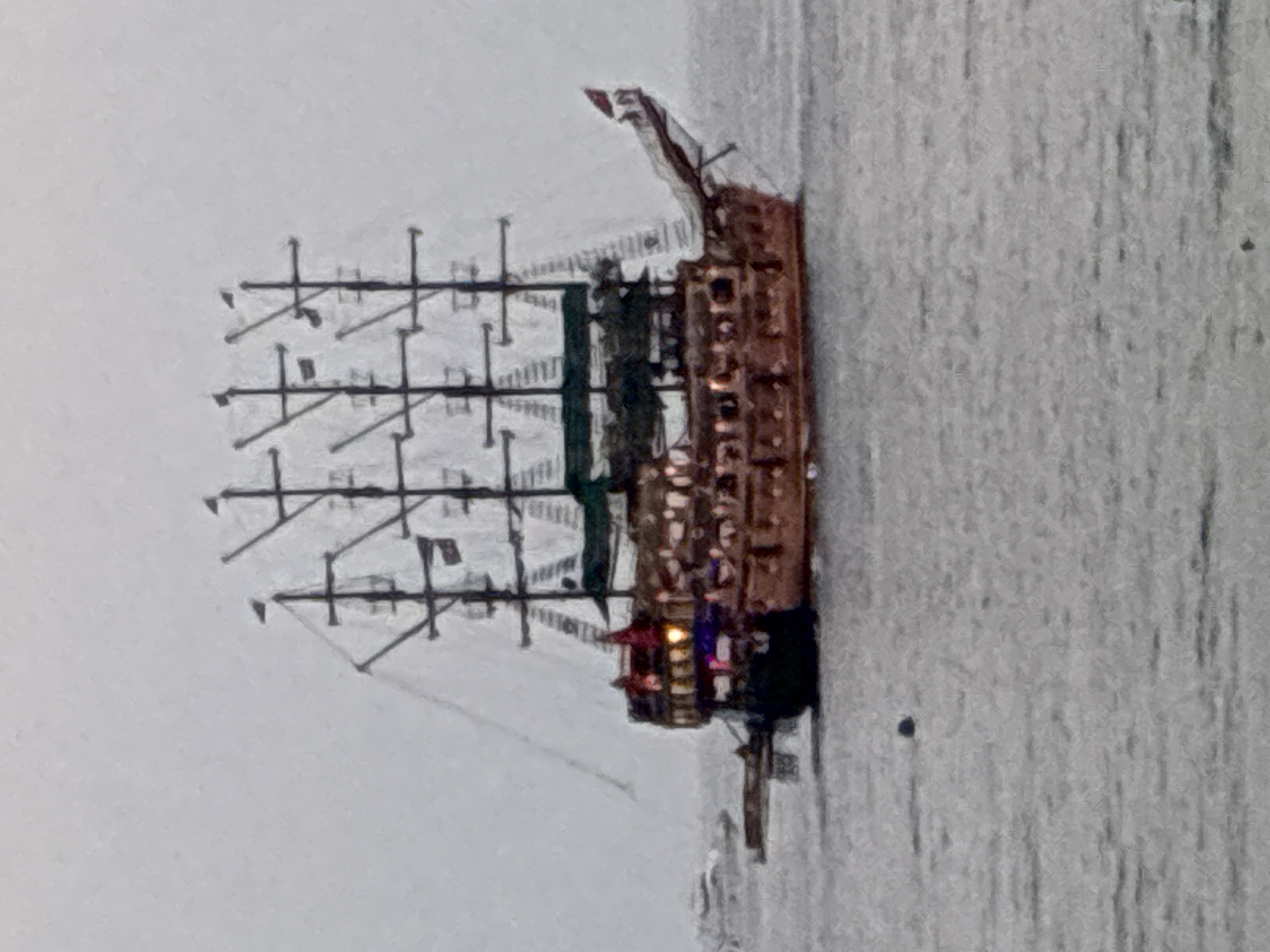 A blurry, overcast image captures a large, wooden, multi-masted pirate ship on the water. The ship has several decks and appears to be illuminated from within. There are flags flying from the masts and the stern. In the distance, on the left side of the frame, another, smaller boat is visible. The water is choppy and grey. The image is taken from a low angle, likely from another boat or the shore. The scene is consistent with the Alanya Marina in Alanya, Turkey, which is known for its tourist-oriented pirate ship excursions.