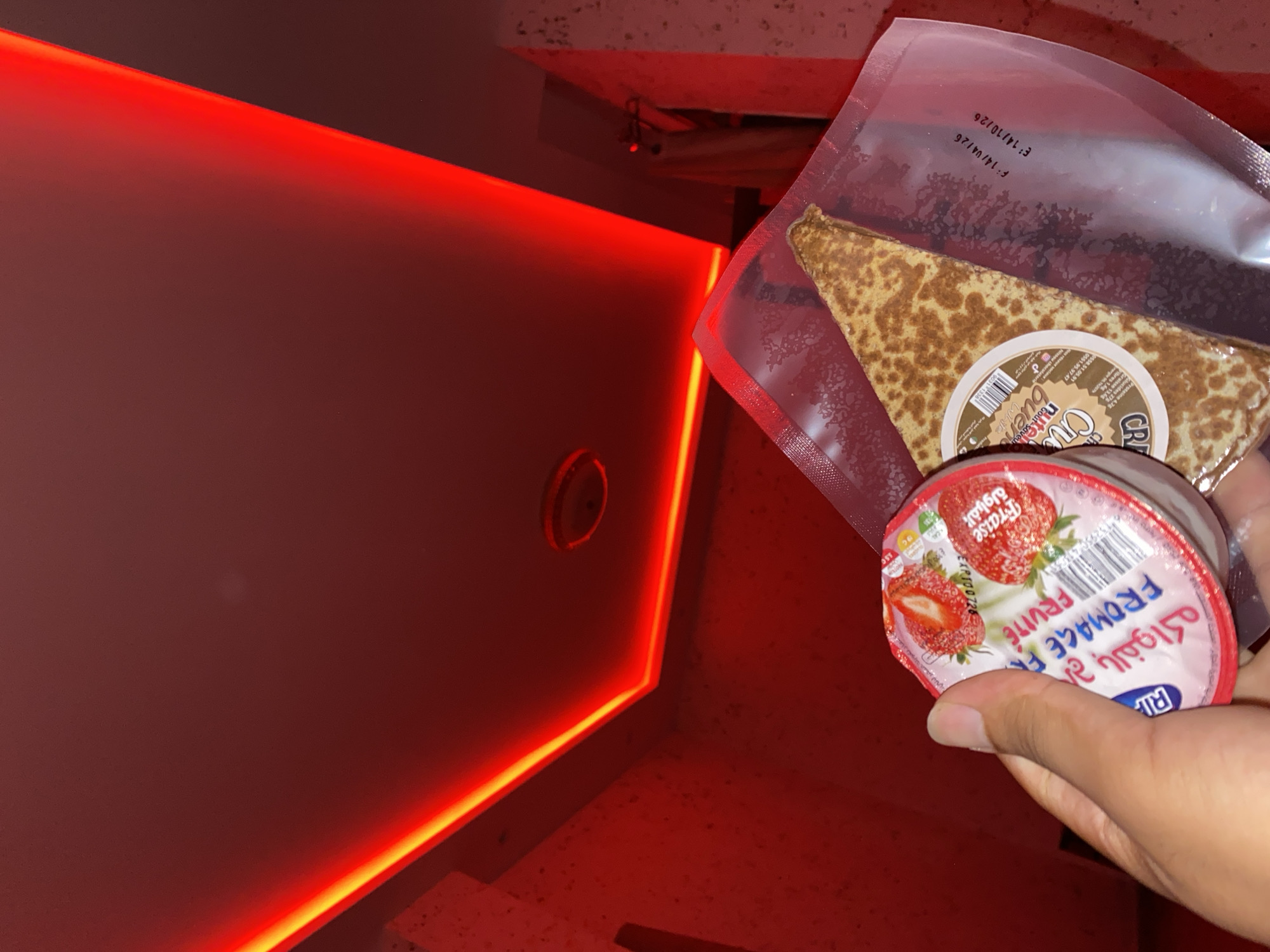 In a dimly lit room with vibrant red and yellow LED strip lighting, a hand holds a small, round container of strawberry yogurt. The yogurt container is white with a pink lid adorned with images of strawberries and text in Arabic and French, including "Fromage F**ruit**e" and "BALOUKH". To the right of the yogurt, a folded crepe is visible inside a clear plastic package. The crepe appears to be golden brown. The background is a textured red wall, and a section of a ceiling with a circular fixture is visible at the top of the frame. The presence of the food items suggests a residential setting or a place where food is consumed. The lighting creates a dramatic and moody atmosphere. The location, Baraki, Algeria, is not visually depicted in the image itself, but is provided as context.