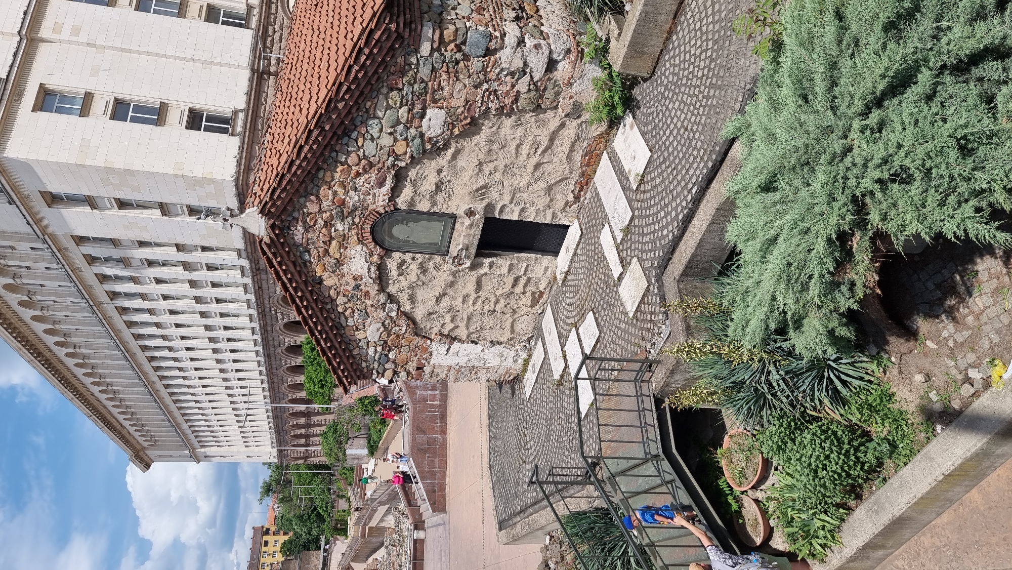 The image shows an elevated view of the Saint George Rotunda in Sofia, Bulgaria, a preserved early Christian church. The building is made of brick, stone, and mortar, with some modern additions. Adjacent to the church, a multi-story white building with repetitive window patterns is visible. A section of Roman ruins is partially exposed, featuring walkways and staircases. Several people are present, walking along the path and observing the ruins. A variety of plants and greenery, including trees, shrubs, and potted plants, add to the scenery. The sky is blue with scattered clouds.