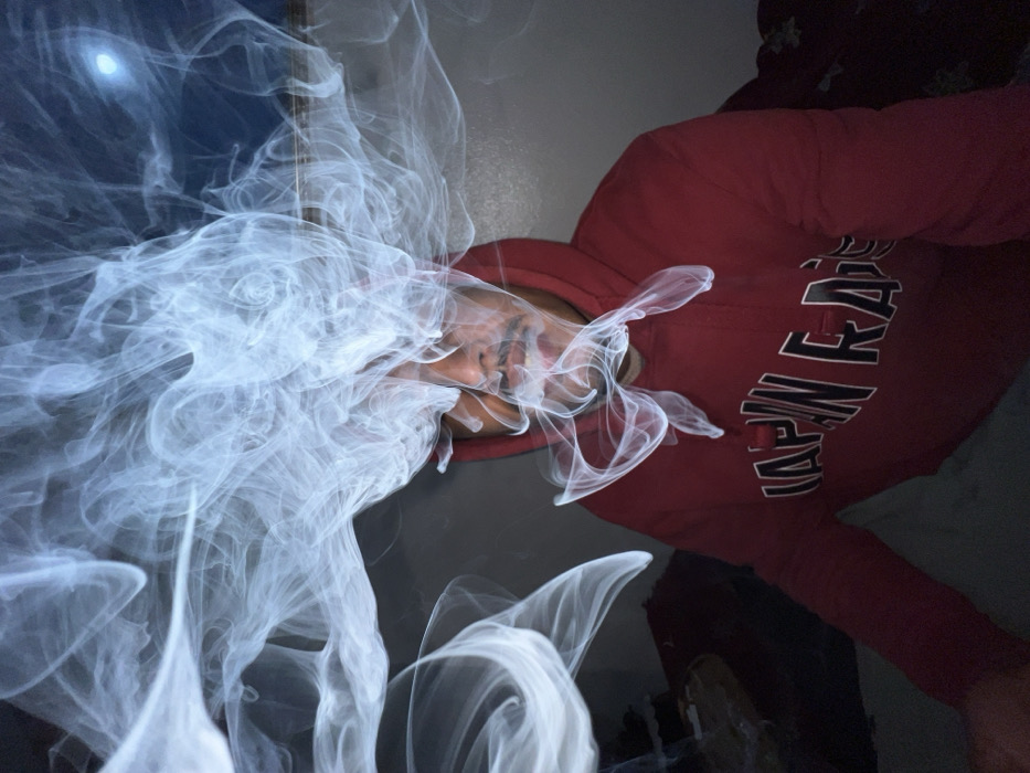 This image depicts a male individual positioned indoors within the city of Tangier, Morocco. The subject is shown from the chest up, wearing a dark red hooded sweatshirt featuring the text "JAPAN RAISED" in white lettering across the chest. He has dark skin and a mustache, displaying a neutral to slightly smirking facial expression. Voluminous, illuminated white smoke intricately swirls around and partially obscures his head and upper torso. The background is dark and largely indistinct, indicating an indoor setting with subdued lighting; a faint circular light source is visible in the upper right quadrant. The smoke appears to actively envelop and surround the individual.