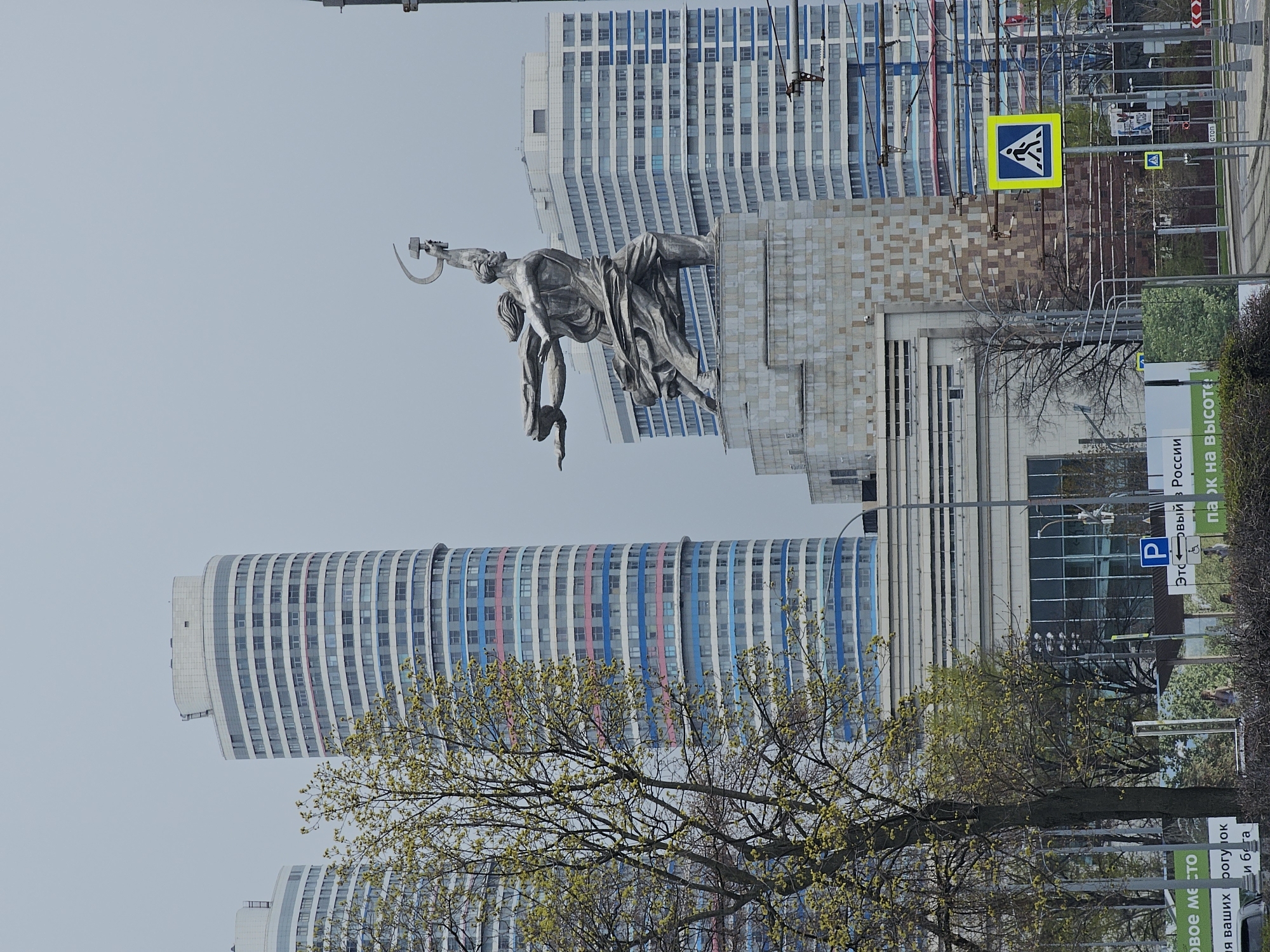 The image depicts the iconic "Worker and Kolkhoz Woman" statue in Moscow, Russia, set against a backdrop of modern high-rise buildings. The statue, a monumental sculpture of a man and woman holding aloft a hammer and sickle, is prominently featured. In the background, two skyscrapers with distinct architectural designs are visible. One has a curved façade with horizontal bands of color, while the other appears more traditional with a grid of windows. A tree with emerging leaves is in the foreground, partially obscuring the buildings. A pedestrian crossing sign is located to the right of the statue. Street signs and a parking symbol are partially visible in the lower right.