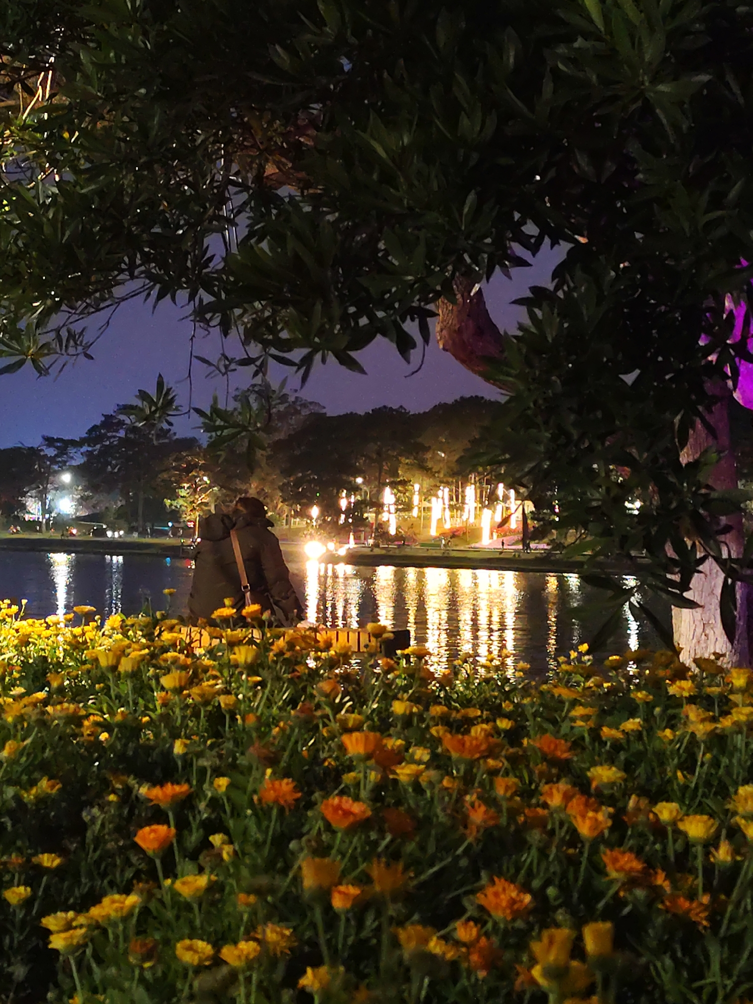 A nighttime shot at Xuan Huong Lake in Da Lat, Vietnam, shows a bed of yellow and orange flowers in the foreground, with the lake reflecting lights in the background. Two people are standing near the lake. Trees frame the scene, with one tree on the right illuminated with purple light. Other trees, some adorned with decorative lights, line the lake's far side. The sky is dark with a hint of twilight.