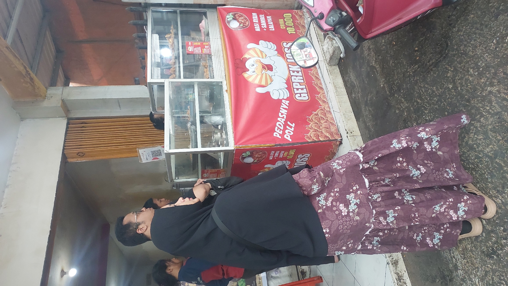 The image shows a street food stall in Malang, Indonesia, selling "Geprek Joss" chicken. In the foreground, a woman in a black hijab and a floral patterned dress stands with her back to the camera, looking towards the stall. She is wearing light-colored platform sandals. Behind her, a man in glasses and a black head covering is also visible, along with other people at the stall. The stall itself is decorated with a vibrant red banner featuring a cartoon chicken giving a thumbs-up and text advertising "Nasi Ayam + Sambal Lalapan Cuma 10.000" (Chicken Rice + Sambal Lalapan Only 10,000). There are glass display cases containing food items, and a motorcycle is parked to the right of the stall. The ground is a dark, weathered asphalt surface. The background shows a building with a yellow accordion-style door.
