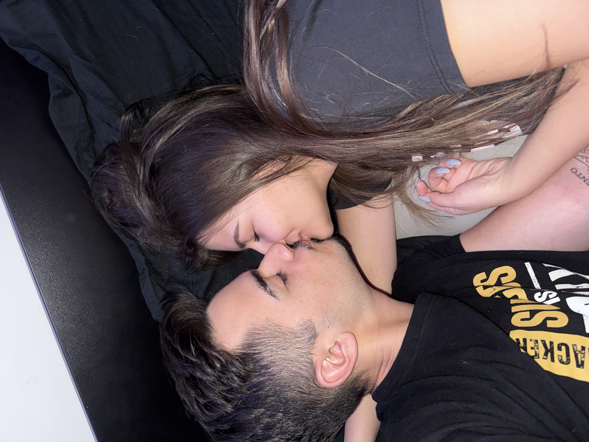 This image depicts an intimate scene featuring an adult male and an adult female engaged in a kiss, positioned closely together in what appears to be an indoor setting, likely a bed or lounge area.
The male, situated on the left, has short dark hair, a faint mustache, and light stubble/beard. His eyes are closed. He is wearing a black short-sleeved t-shirt with partially visible yellow and white text that reads "LACKER" and "SISTERS IS ONLY". His right arm is partially visible behind the female's head.
The female, on the right, has long dark brown hair with lighter reddish-brown highlights, flowing across a dark surface. Her eyes are closed. She is wearing a black short-sleeved t-shirt. Her left hand is visible, featuring light blue nail polish, and appears to be holding or resting near the male's hand/arm. A partial tattoo, reading "ENTO," is visible on her lower left arm or wrist.
Their lips are touching, indicating a kiss. The immediate background consists primarily of dark, possibly black, fabric (likely bedding). In the upper left corner, there is a distinct horizontal line separating a white surface from a dark, textured surface, with a thin blue/purple line running along the edge. The lighting is soft and appears to be artificial.
The specific geographical location, including the city of Sofia, Bulgaria, cannot be determined from the visual evidence within the image.