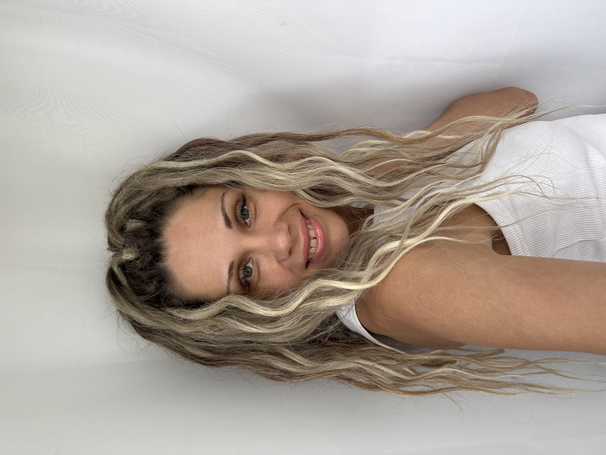 A medium shot captures a woman with blonde hair styled in loose waves and dreadlocks against a white backdrop. She is positioned in a corner, with the white wall curving behind her. The woman is looking directly at the camera and smiling. She is wearing a white tank top. The image is a portrait, and no specific location in Rusanovo, Russia is discernible.