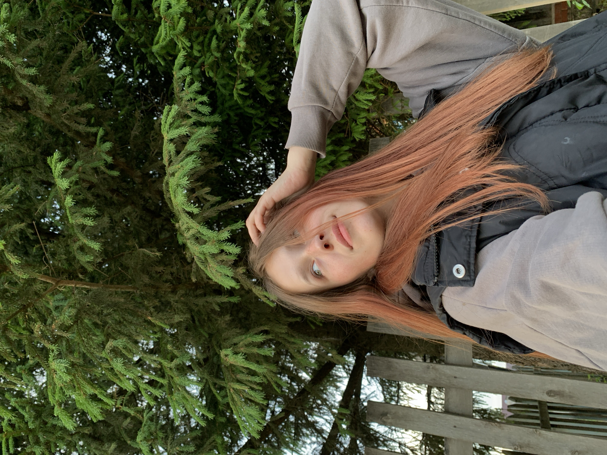 A young, fair-skinned person with long, light orange hair is positioned in front of a dense evergreen tree. The person is wearing a light gray sweatshirt or hoodie and a black vest. They have their hand near their forehead, possibly touching their hair. The background also reveals a wooden fence or structure, suggesting an outdoor location, likely a park or garden. The photo is taken from a low angle, looking up. The location is Фоки, Russia.