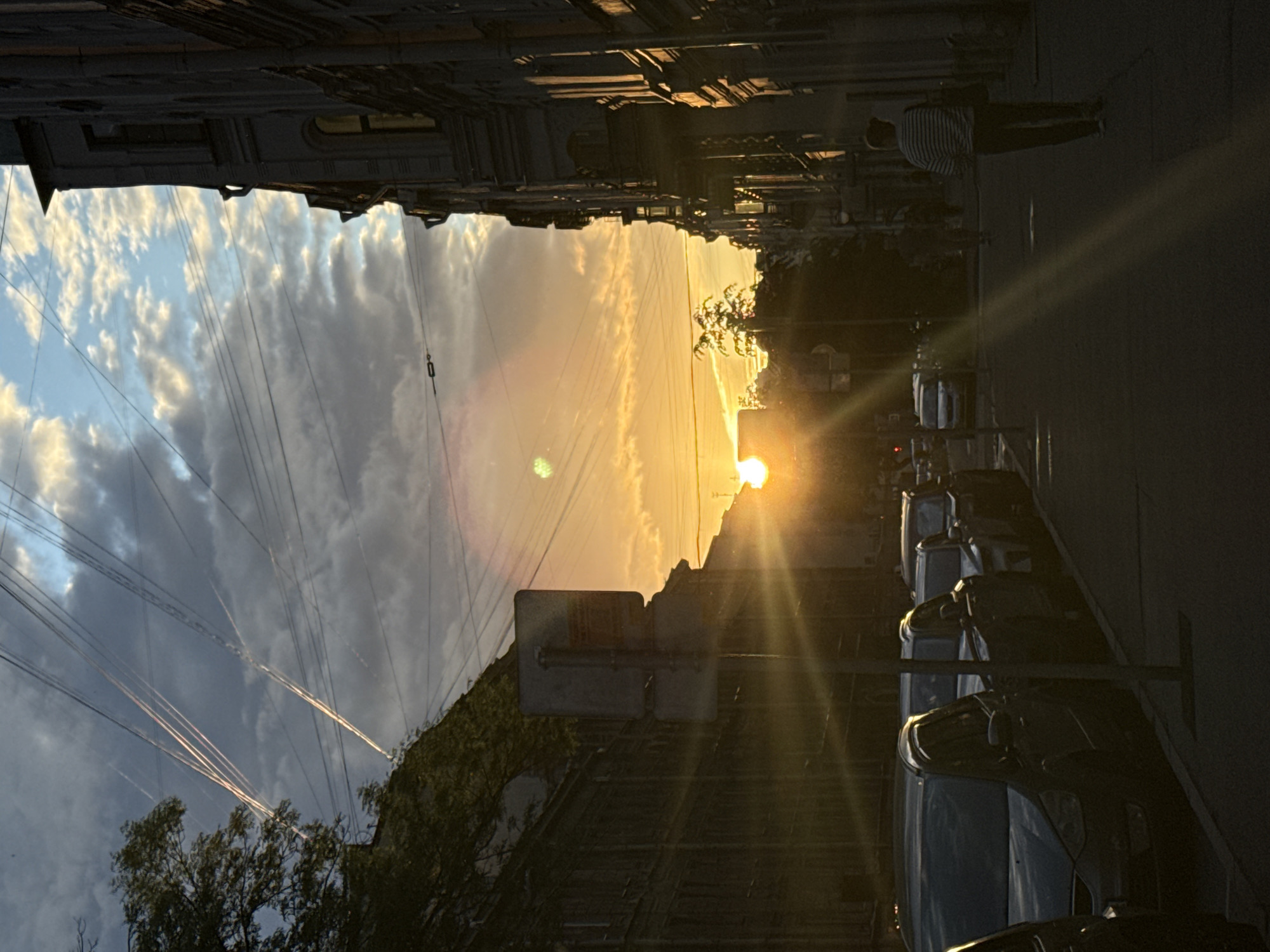The image depicts a street scene in Saint Petersburg, Russia, during sunset. The sky is a mix of dramatic clouds and a bright orange glow from the setting sun, with visible contrails from aircraft. Buildings, characteristic of Saint Petersburg's architecture, line the street on both sides. On the left, a multi-story building with trees in front is visible, while on the right, a grander, ornate building is silhouetted against the bright sky. Several cars are parked along the street, mostly dark sedans and SUVs. A street sign stands on the left side of the road. In the distance, a few individuals are walking, with one person in the foreground on the right wearing a striped shirt. The overall lighting suggests the golden hour, with strong sun rays creating lens flare and illuminating parts of the scene.