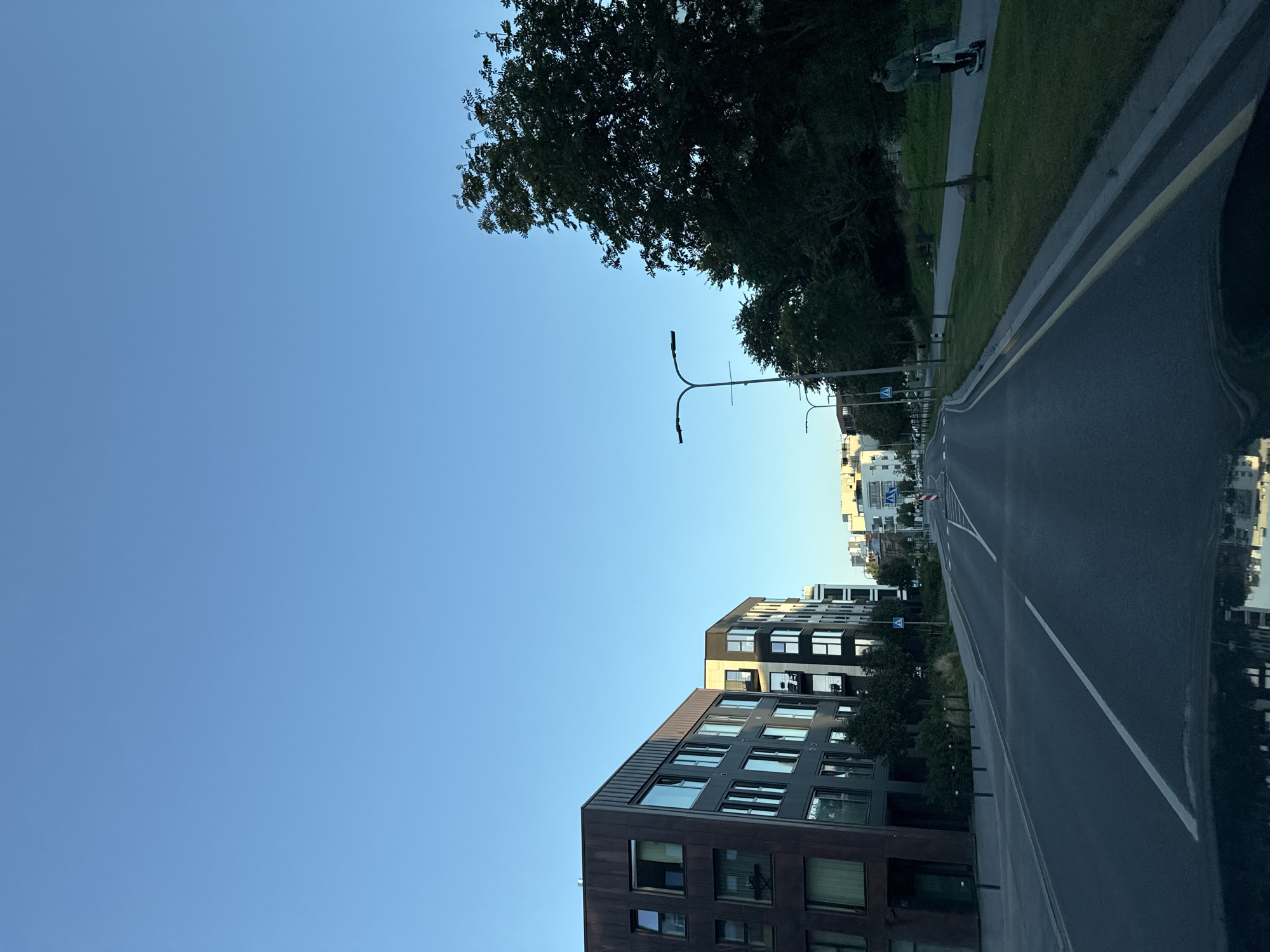 The image shows a street scene in Tallinn, Estonia, likely in a modern urban district. On the left, a contemporary apartment building with dark brown and gray facades and large windows is visible. Further down the street, other similar residential buildings line the road. The street itself is paved and has lane markings. A grassy area with trees and streetlights runs along the right side of the road. A person is riding an electric scooter on a paved path in this grassy area. The sky is clear and blue, indicating daylight. The overall impression is a clean, well-maintained urban environment with a mix of residential architecture and green spaces.