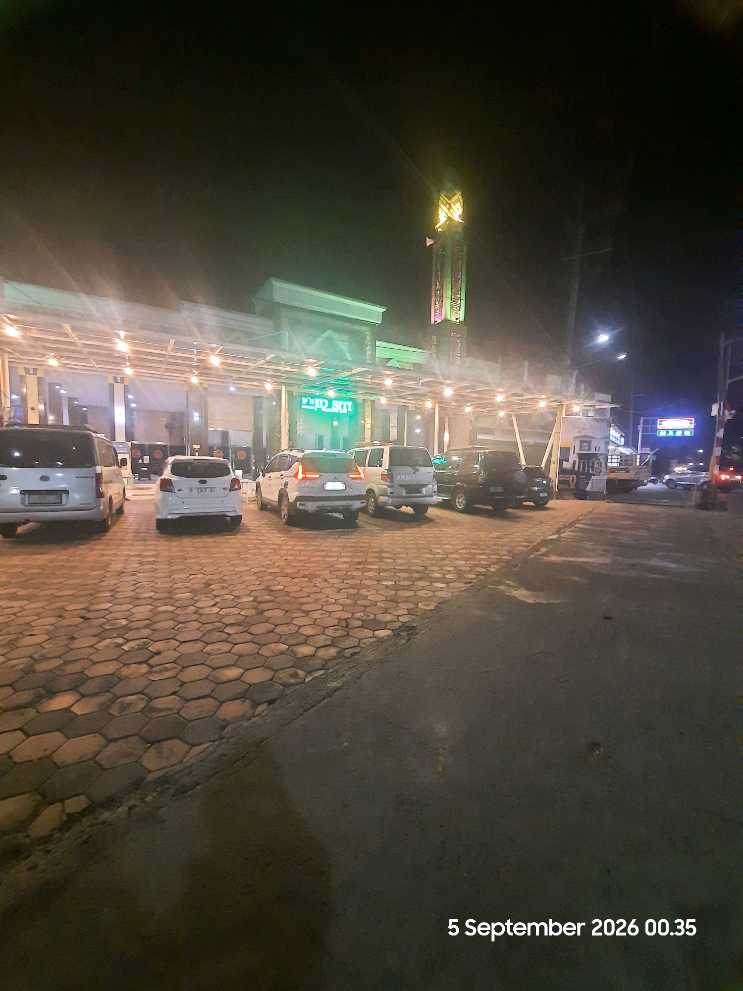 The image shows a night scene in Sukokerto, Indonesia, with a mosque prominent in the background. The mosque features a tall, illuminated minaret with green and yellow lights. In front of the mosque, there is a covered outdoor area with lights illuminating a parking lot. Several cars are parked in the lot, including a white van, a white hatchback, and a white SUV. The ground is paved with hexagonal bricks. The overall impression is of a well-lit public space at night. The date "5 September 2026 00.35" is displayed in the lower right corner.