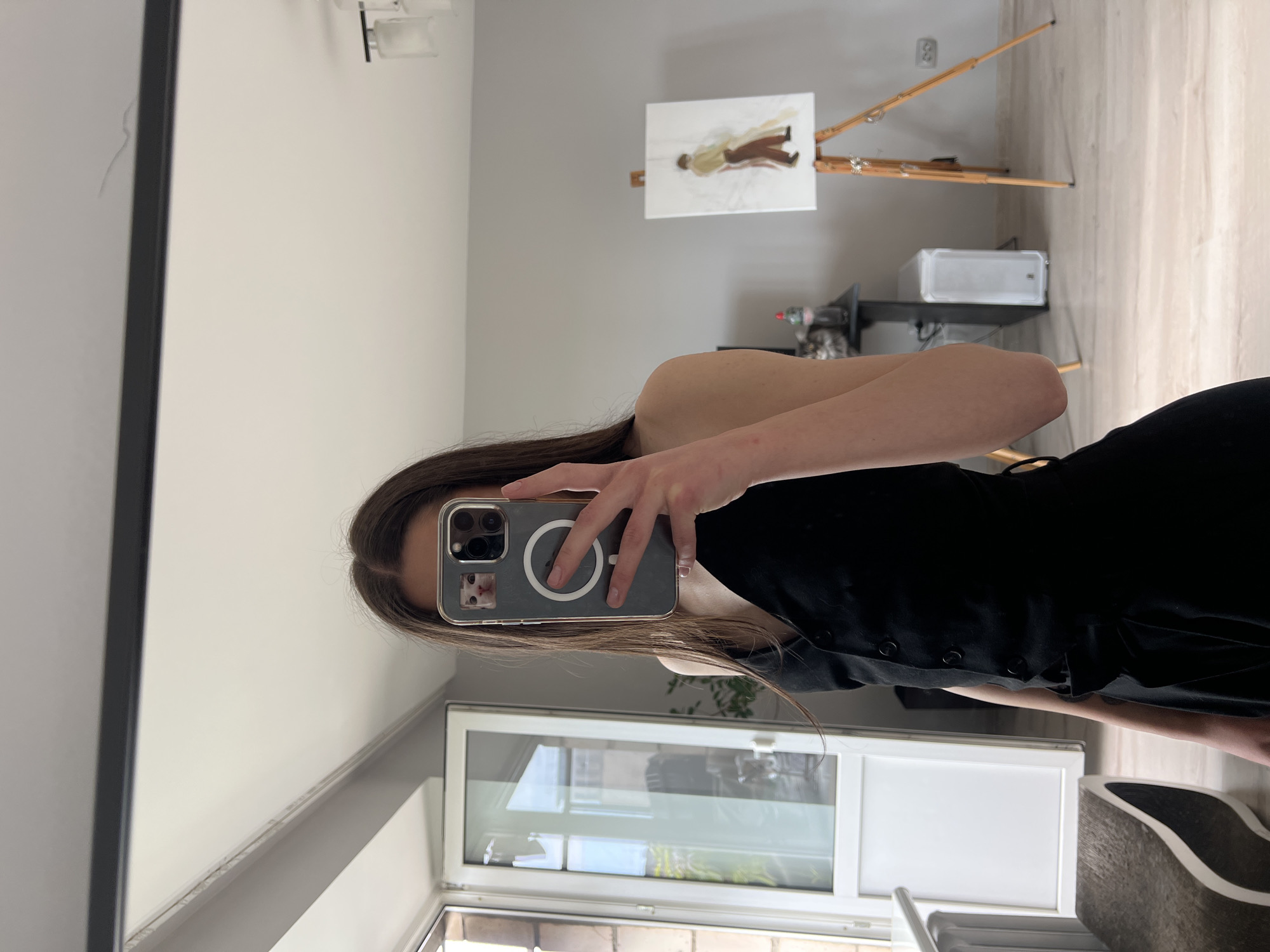 A person with long brown hair and wearing a black dress is holding a phone in front of their face, presumably taking a selfie in a mirror. The phone has a clear case with a design of a cat in the corner. Behind the person, there's a window and the outline of a building visible through the glass.
In the background reflected in the mirror is a wall with a painting on a stand. A computer tower and other objects are on a shelf. The photograph appears to have been taken indoors, with artificial lighting, though the location cannot be determined as Ternopil. Ukraine from this photo.
