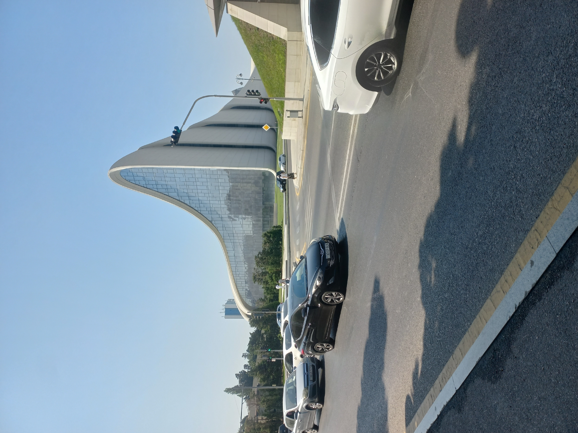 The image depicts the Heydar Aliyev Center in Baku, Azerbaijan, on a clear, sunny day. The iconic, wave-like architecture of the center dominates the background, its white facade contrasting with the blue sky and the large glass panels. In the foreground, a multi-lane asphalt road is visible with several cars. A black sedan with license plate "90-XF-906" is in the right lane, followed by a silver sedan with license plate "99-UV-506". A white sedan is partially visible in the far right lane, and another white car is in the background. Pedestrians are seen walking on a sidewalk near the building and across a crosswalk. Traffic lights and street lamps are also present. The scene suggests a bustling urban environment with modern architecture and active traffic.