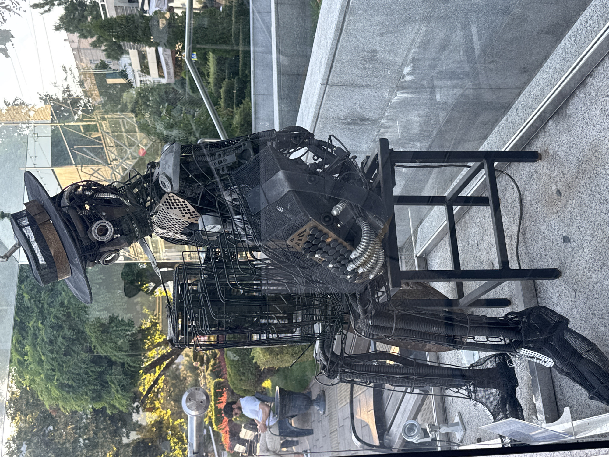 This image depicts a street art sculpture of a steampunk-style robot playing an accordion, located in the city of Rostov-on-Don, Russia. The robot is crafted from scrap metal, featuring gears, pipes, and mesh. It is seated on a black metal stool. In the background, a man in a white t-shirt and light-colored pants is sitting on a bench, looking at his phone. The environment appears to be an outdoor public space with greenery, paved walkways, and modern architecture reflected in a glass barrier. The lighting suggests it is daytime, possibly late afternoon due to the warm tones of the sunlight.