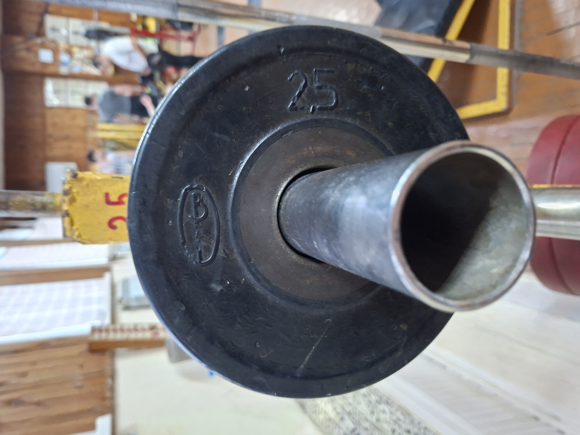 A close-up, eye-level, indoor shot focuses on a black, circular weight plate with the number "25" imprinted on it, attached to a metal barbell. The plate has a circular opening in the center where the barbell passes through, and a metallic ring around the opening. The background is blurred, showing what appears to be a gym with wooden walls and flooring. Several out-of-focus individuals are visible in the background, engaged in various activities. The scene is brightly lit. The image is undated and its location is unknown.