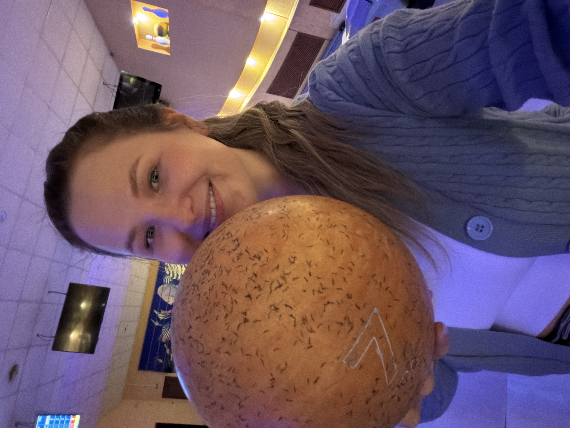 A woman is holding a bowling ball in a bowling alley. She is smiling and looking at the camera. The bowling ball is orange with a white arrow on it. She is wearing a light blue cardigan sweater over a white top. The bowling alley has TV screens and a ceiling installation. The city of Chaikovsky, Russia is not identifiable in this image.
