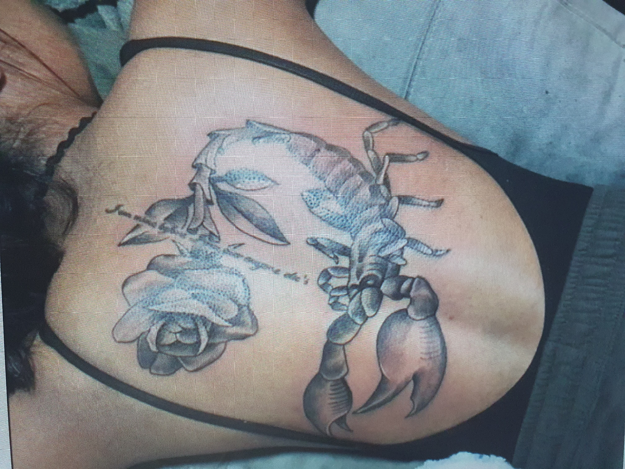 The image shows a tattoo of a scorpion intertwined with a rose on the back of a person. The tattoo is rendered in black and grey with shading. The person is wearing a dark bra or tank top. The background is out of focus and appears to be an indoor setting. The image is a photograph taken from an angle looking down at the person's back. There is no information in the image to indicate the location in Tashkent, Uzbekistan.