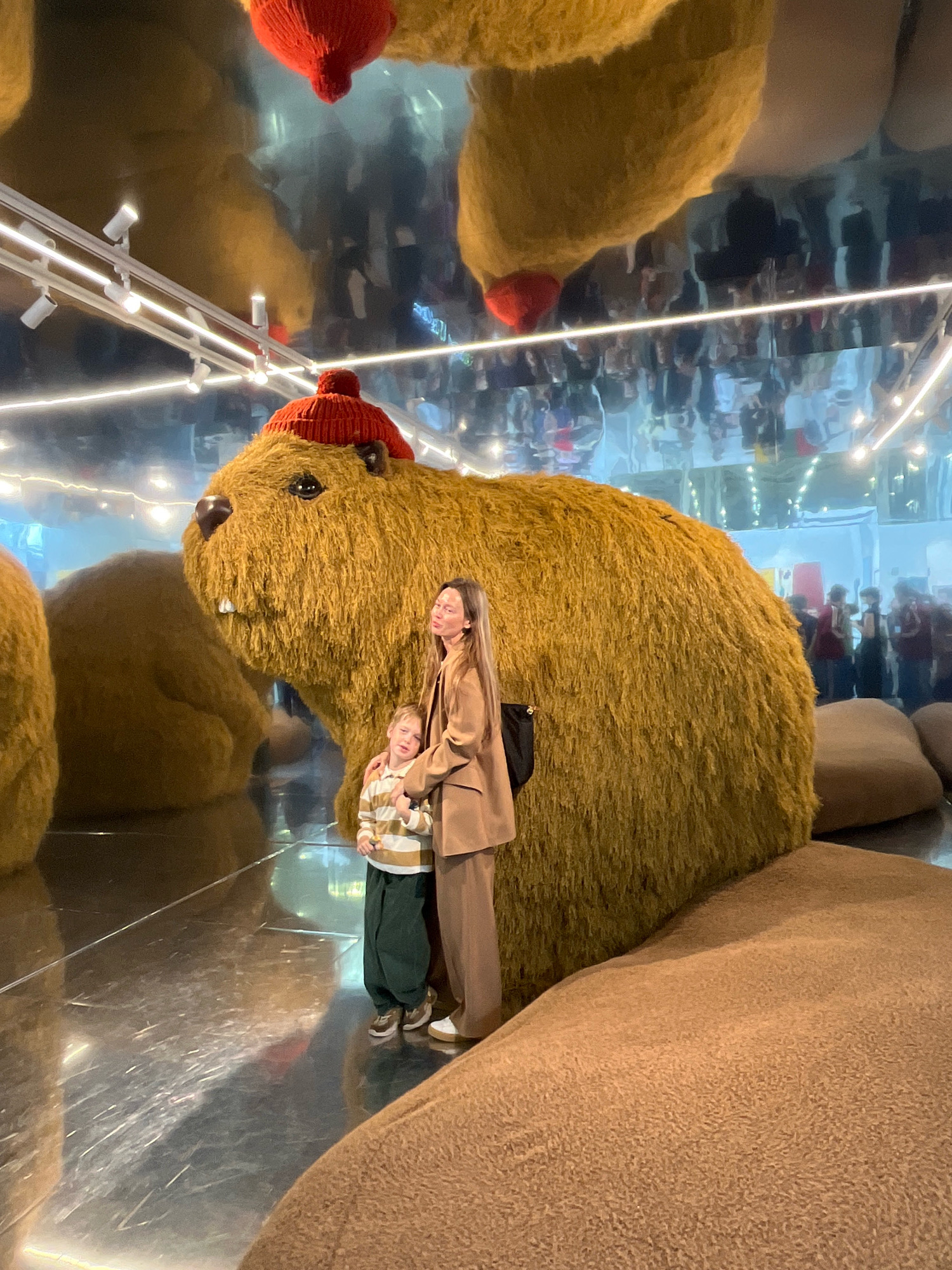 A woman and a child stand next to a giant, fuzzy, tan capybara sculpture wearing a red knitted hat. The sculpture has large, bulbous forms that appear to be part of a larger installation. The environment features a highly reflective floor and ceiling, creating distorted reflections of the sculpture and other elements in the space, including lights and people. The overall scene suggests an indoor exhibition or artistic installation. The location in Khimki, Russia, is not evident from the visual information provided in the image.