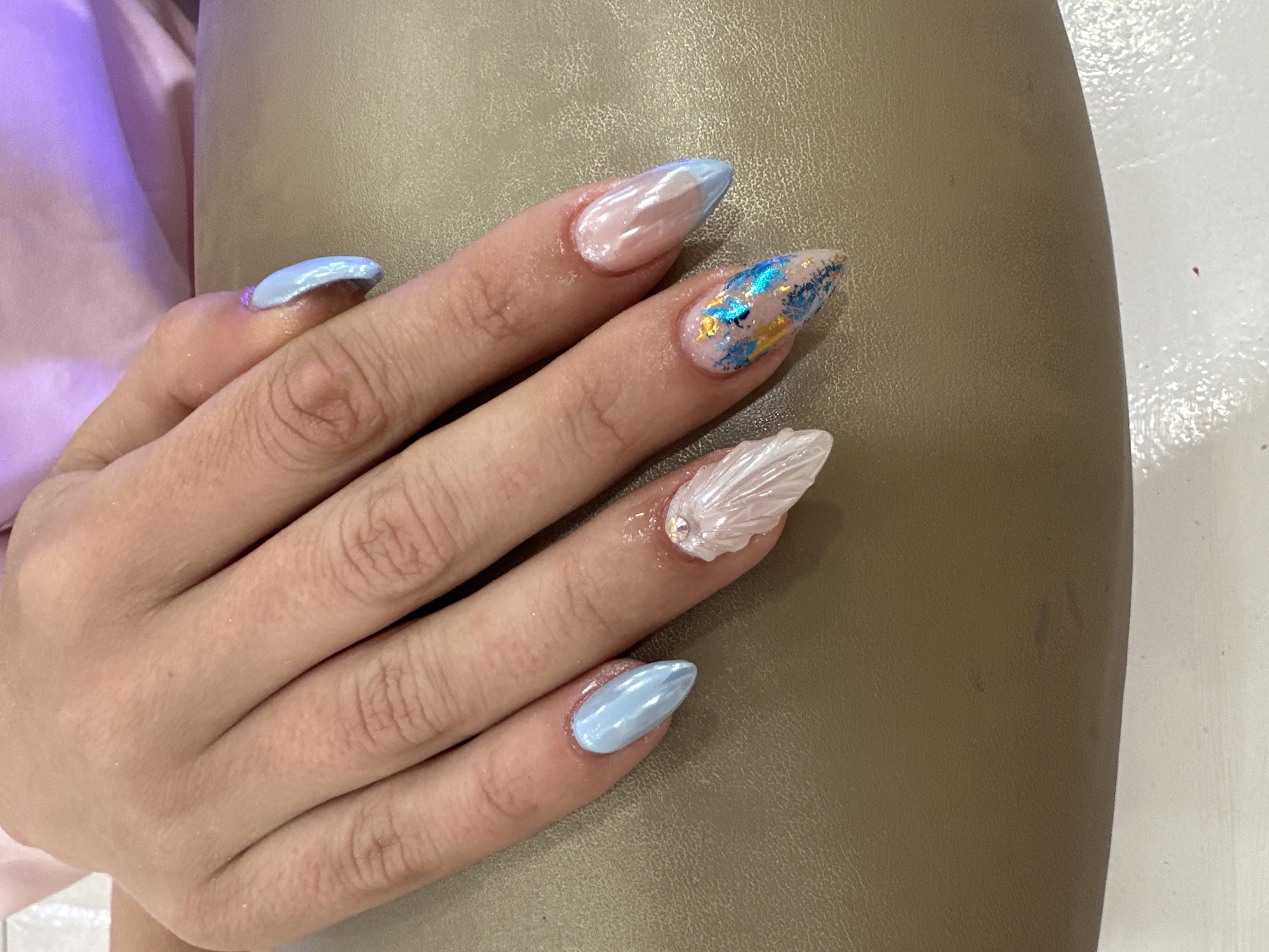A close-up shot of a person's left hand, displaying a set of almond-shaped acrylic nails with various designs. From the index finger to the pinky, the nails feature:
1. A classic French tip design with a light blue tip.
2. Glittering blue and gold flakes over a pale pink base.
3. A sculptural design resembling a shell, adorned with a small gemstone near the base.
4. Light blue polish with a metallic sheen.
The hand is resting on a smooth, khaki-colored surface with light purple textile visible in the background. The environment appears to be indoors, with hints of white or light-colored surfaces in the background. The image does not reveal any discernible landmarks or indicators that would pinpoint its location to Sofia, Bulgaria.