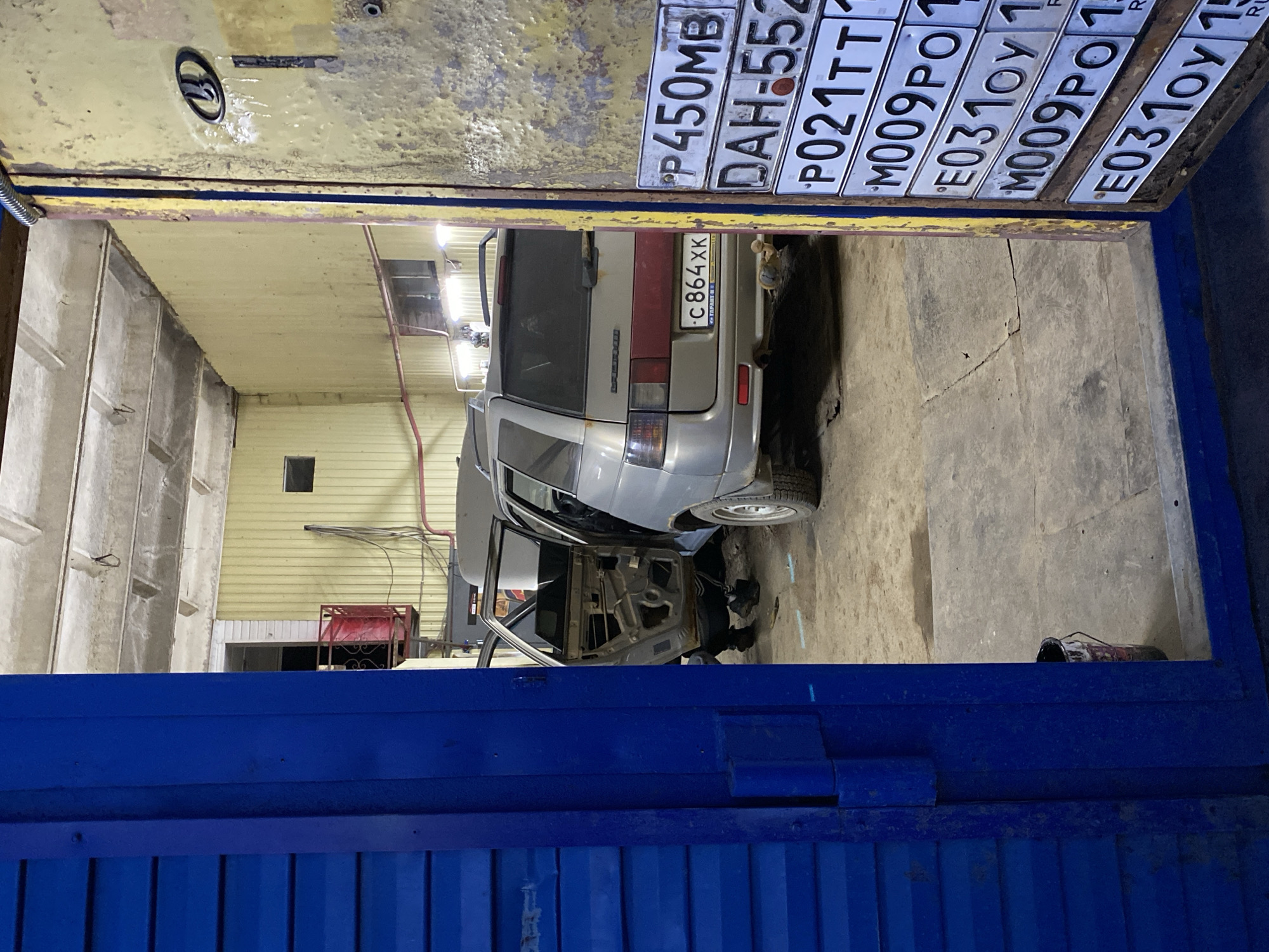 The image captures a garage scene, likely in Chaikovsky, Russia, based on the Russian license plates. Two cars are present. One is a silver station wagon with visible damage, its rear end closest to the viewer, displaying a license plate reading "c864 XK". The other car's door frame is visible, indicating a project or repair work in progress.
The garage itself is cluttered with a variety of items, including a rack and tools scattered around. Notably, a collection of license plates is mounted on the garage door, bearing codes such as "P450MB", "DAH-552", "P021TT", "M009PO", and "E031ΟΥ 1." The garage door is a combination of blue and yellow, with the blue section partially open at the bottom. The walls of the garage are a light, faded color. The lighting is provided by fluorescent fixtures on the ceiling.