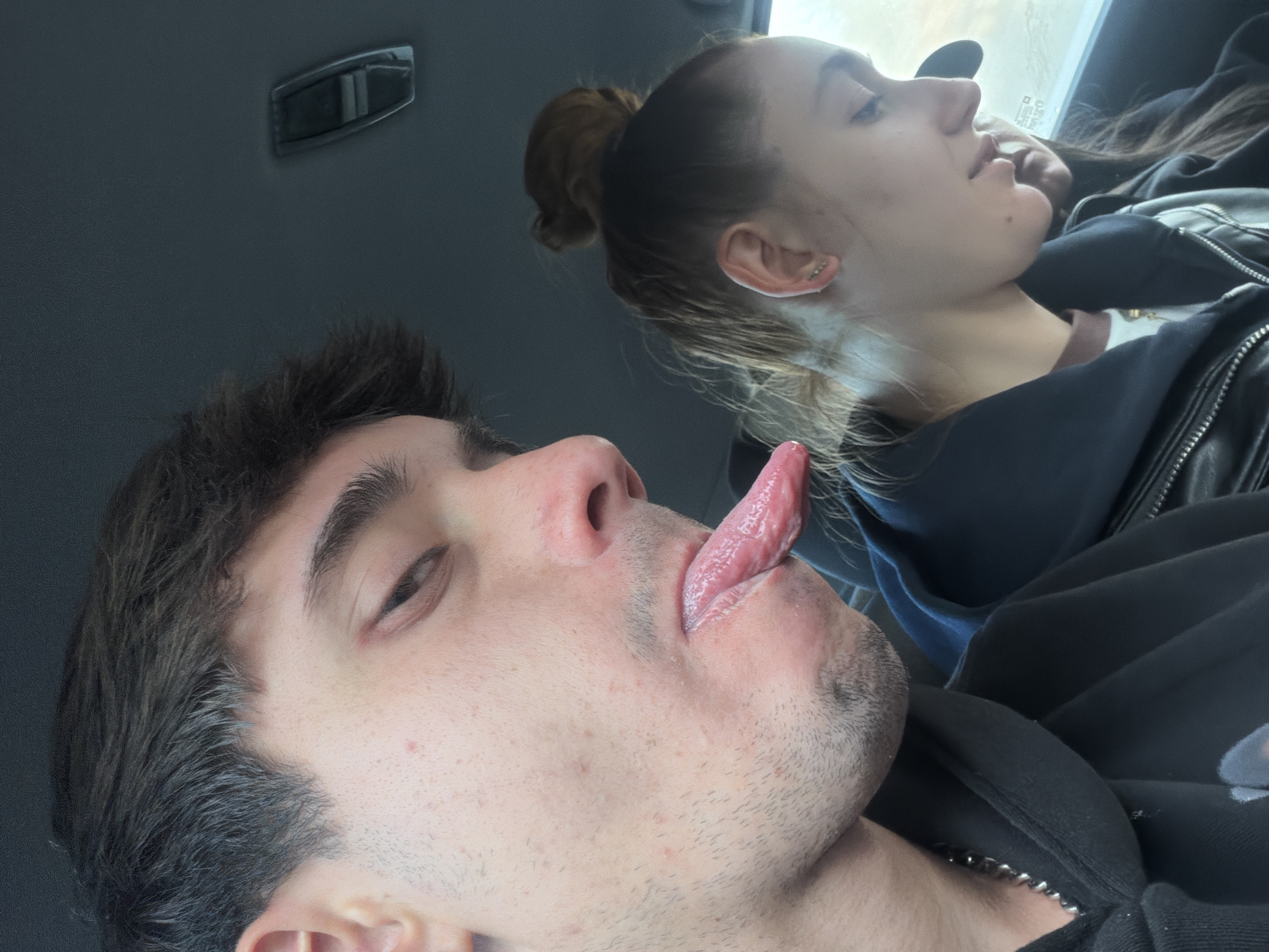 Two individuals, a male in the foreground and a female in the background, are captured inside a vehicle in Staro Selo, Bulgaria. The image is taken from a low angle, looking upwards, and is rotated approximately 90 degrees clockwise.
The male subject, positioned in the left foreground, is seen from a profile/three-quarter view, playfully sticking his tongue out. He has dark, wavy hair, visible stubble on his face, and wears a dark, zippered hoodie or jacket, with a silver-colored chain necklace visible.
The female subject, behind and to the right of the male, appears to be resting or sleeping with her eyes closed. Her light brown hair is pulled back into a bun. Her visible ear features multiple piercings, including studs and a barbell. She is wearing a dark jacket, possibly leather, over a darker top, with a lighter-colored collar visible underneath.
The setting is inside a car, with a dark gray headliner and a grab handle visible overhead. Bright light streaming through a window suggests it is daytime. The view outside the window is blurred, indicating either motion or a shallow depth of field focusing on the subjects. A faint, illegible white graphic or text is partially visible on the male's dark clothing.