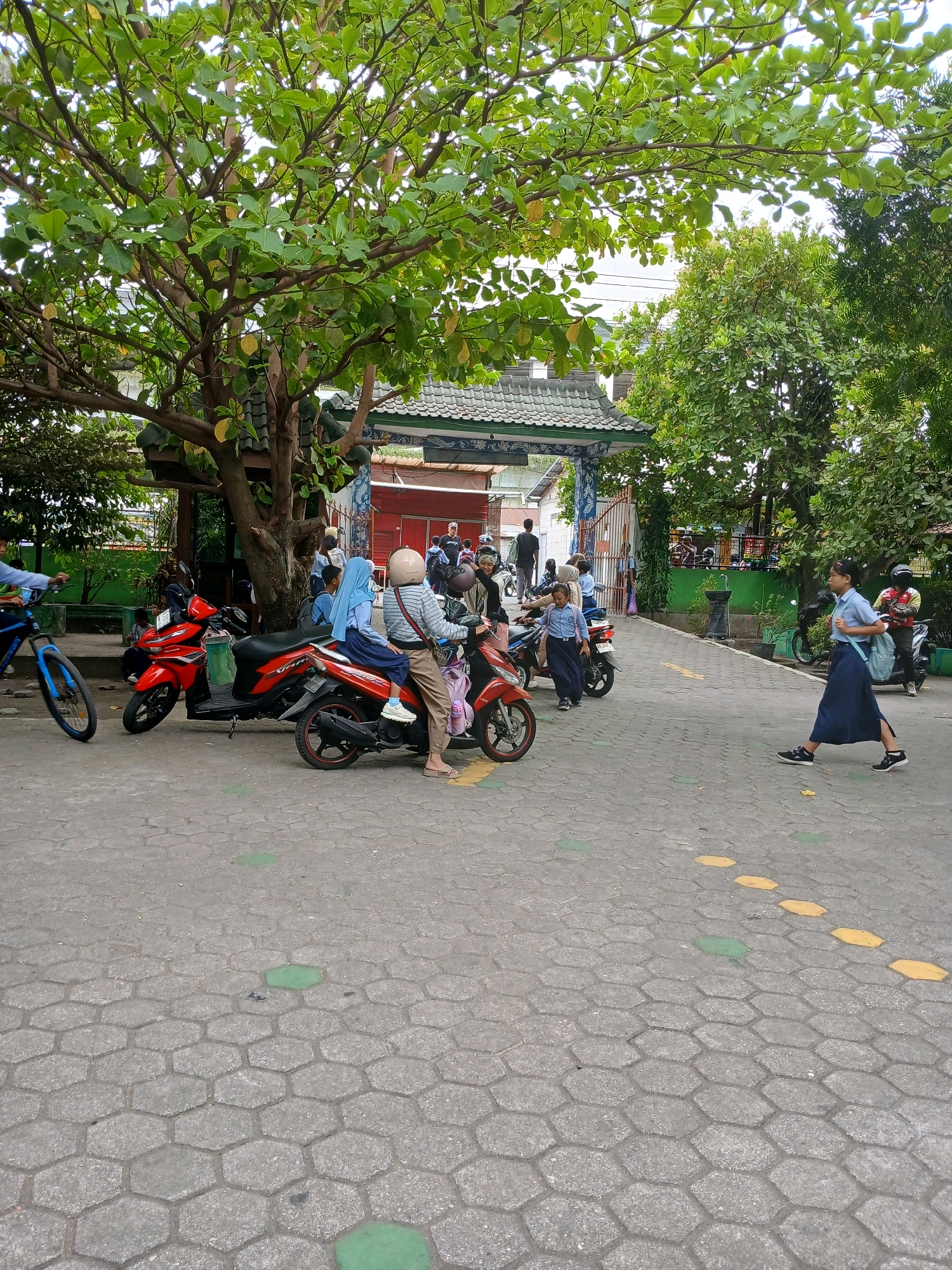 The image depicts an outdoor scene in Depok, Indonesia, likely near a school or public gathering area. In the foreground, a paved ground surface made of hexagonal blocks is visible, marked with yellow and green painted shapes. A young girl wearing a blue uniform and skirt, with a backpack, is walking across the paved area from right to left. Behind her, further to the right, is a motorcycle with a person in a red shirt and helmet.
The middle ground features several parked motorcycles, including a red one with a rider wearing a light-colored helmet and a blue hijab. Another person in a similar blue uniform is seated behind the rider on this motorcycle. A person on a blue bicycle is moving from left to right in the left-middle portion of the frame. More people, some in uniforms and some in casual attire, are visible in the background, near a gate structure with a tiled roof.
The background is dominated by lush green trees with broad leaves, suggesting a tropical or subtropical climate. The overall setting appears to be a moderately busy public space, possibly the entrance or exit of a school, with a mix of pedestrians and motorcycles.
