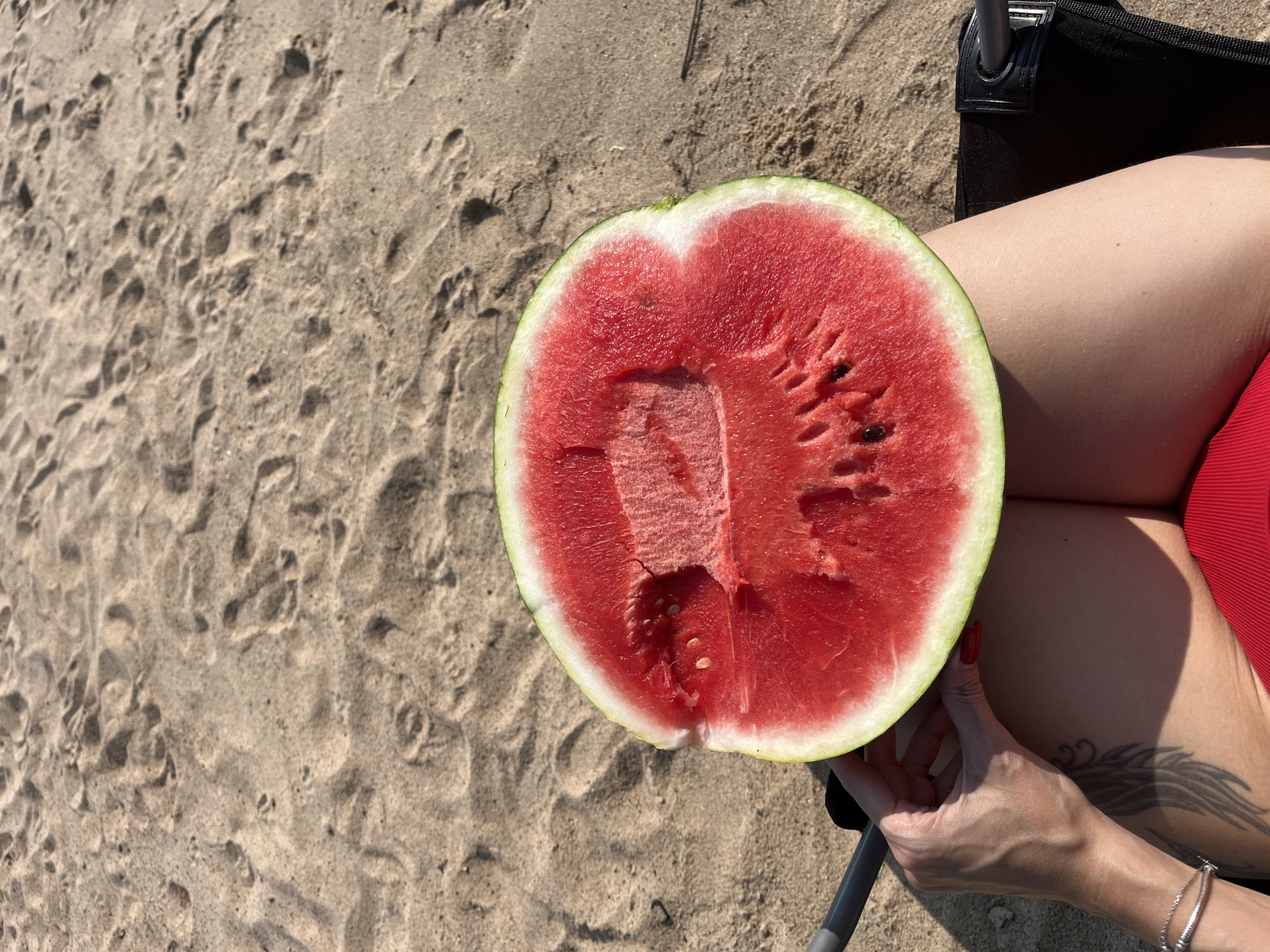 A person is sitting on a beach in Nizhny Novgorod, Russia, with a half-eaten watermelon on their lap. The person is wearing a red swimsuit and has a tattoo on their left arm. The beach is sandy, with footprints visible in the sand. The weather appears to be sunny.