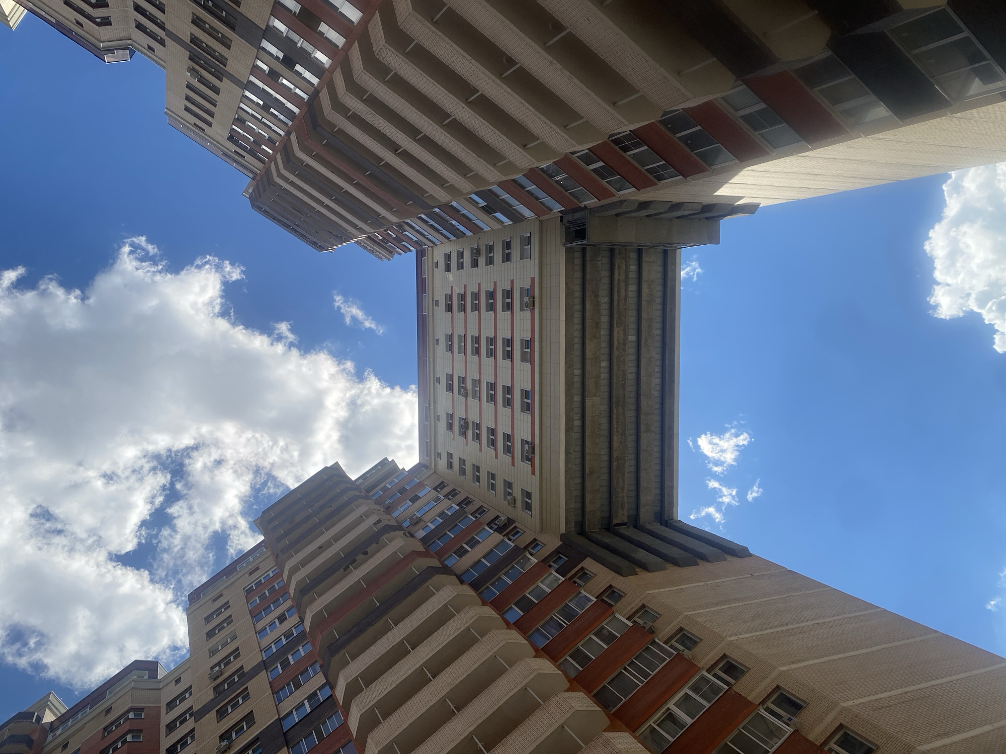 The image captures a low-angle view of modern residential buildings under a bright blue sky with scattered white clouds. The architecture features a mix of light-colored brickwork and darker accent panels, with numerous balconies and windows. One building is positioned directly above another, creating a dramatic visual effect, and appears to be connected by an elevated walkway or bridge. The scene is oriented such that the sky fills a significant portion of the frame, suggesting a clear, sunny day. The perspective is unusual, looking upwards from the ground or a lower vantage point, emphasizing the scale and height of the structures. No people or specific events are depicted. The time of day appears to be daytime, likely late morning or early afternoon, given the bright illumination and the quality of the light. The location is identified as Kotelniki, Russia.