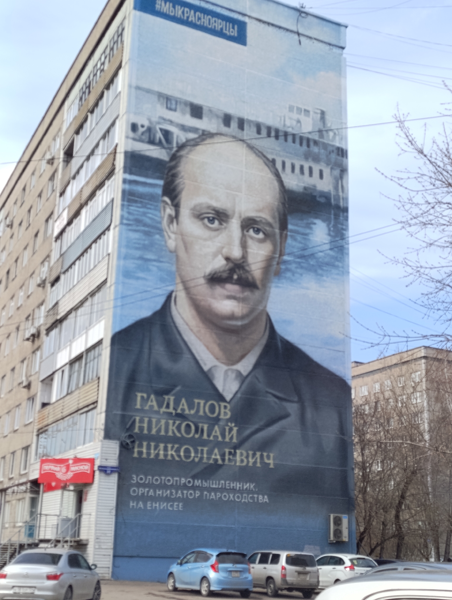 The image presents an urban street scene in Krasnoyarsk, Russia, featuring a large mural on the side of a multi-story building. The mural prominently displays a portrait of a man with a mustache, identified by the text as Gadalov Nikolay Nikolaevich. Behind his head, a depiction of a river, likely the Yenisei, with a steamboat is visible.
Above the portrait, the mural includes the Cyrillic hashtag "#МЫКРАСНОЯРЦЫ" (which translates to "#WEAREKRASNOYARSKCITIZENS"). Below the man's name, text in Russian describes his accomplishments: "ЗОЛОТОПРОМЫШЛЕННИК, ОРГАНИЗАТОР ПАРОХОДСТВА НА ЕНИСЕЕ" (meaning "GOLD INDUSTRIALIST, ORGANIZER OF STEAMSHIP NAVIGATION ON THE YENISEI").
To the left of the mural, another multi-story residential building with numerous windows and balconies is visible. At street level, several cars are parked, including a silver sedan and a blue hatchback. A shop with a red sign, partially obscured but appearing to read "ПЕРВЫЙ ВИННОЙ" (First Wine Shop), is located at the ground floor of the adjacent building. The scene is illuminated by daylight under a clear, bright sky, with bare tree branches on the right side of the frame, suggesting a late winter or early spring setting. Power lines traverse the upper portion of the sky. An air conditioning unit is mounted on the mural-covered building near the bottom. No specific activity or event is taking place; it's a static view of the urban environment.