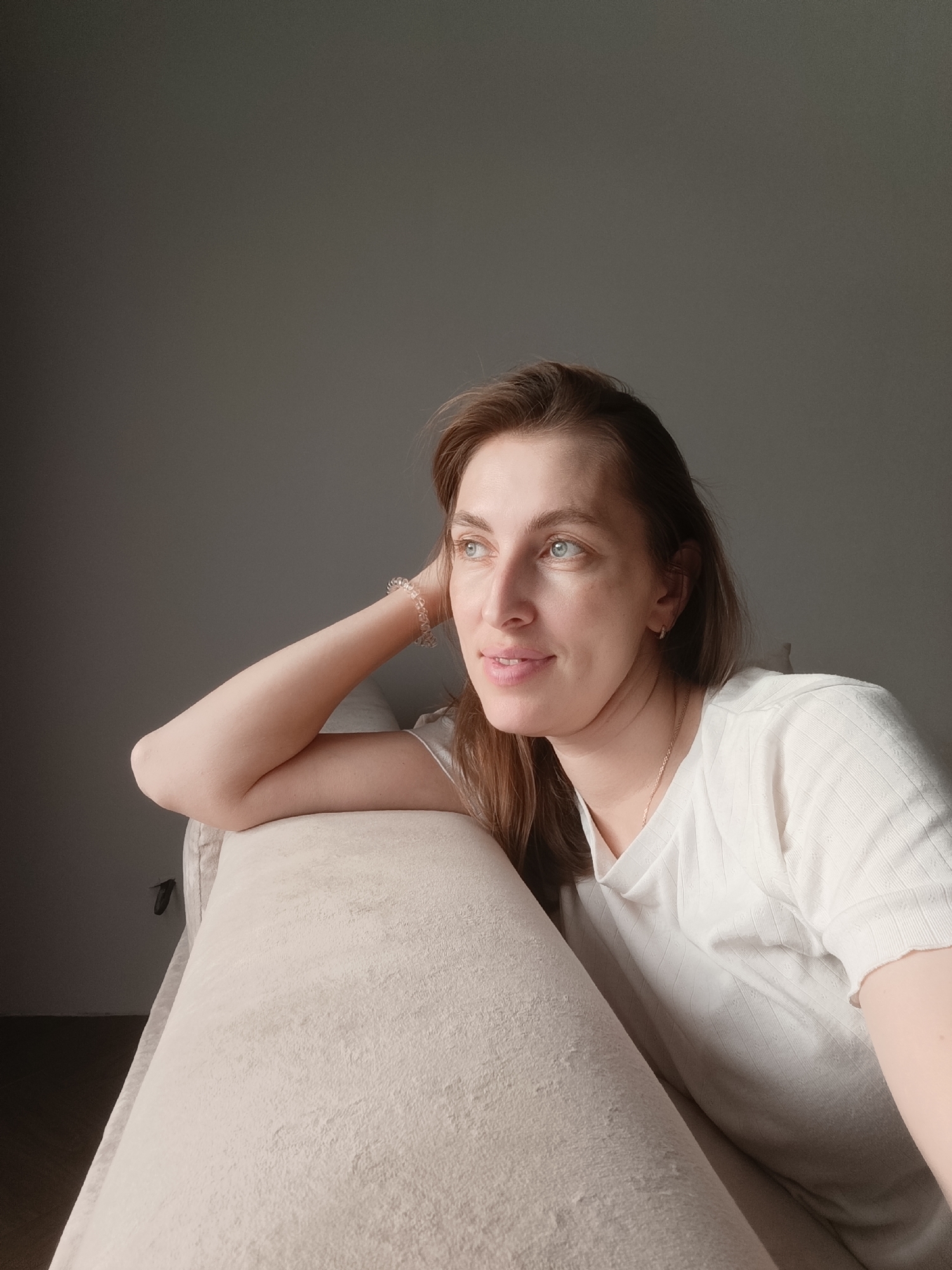 A woman is sitting on a beige sofa, leaning her head on her right arm which is resting on the back of the sofa. She has long brown hair and blue eyes. She is wearing a white t-shirt and a bracelet on her left wrist. The background is a plain gray wall. The image appears to be taken indoors. The setting is not definitively identifiable as being in the city of Chaikovsky, Russia, nor is there any specific information in the visual data that would place it there.