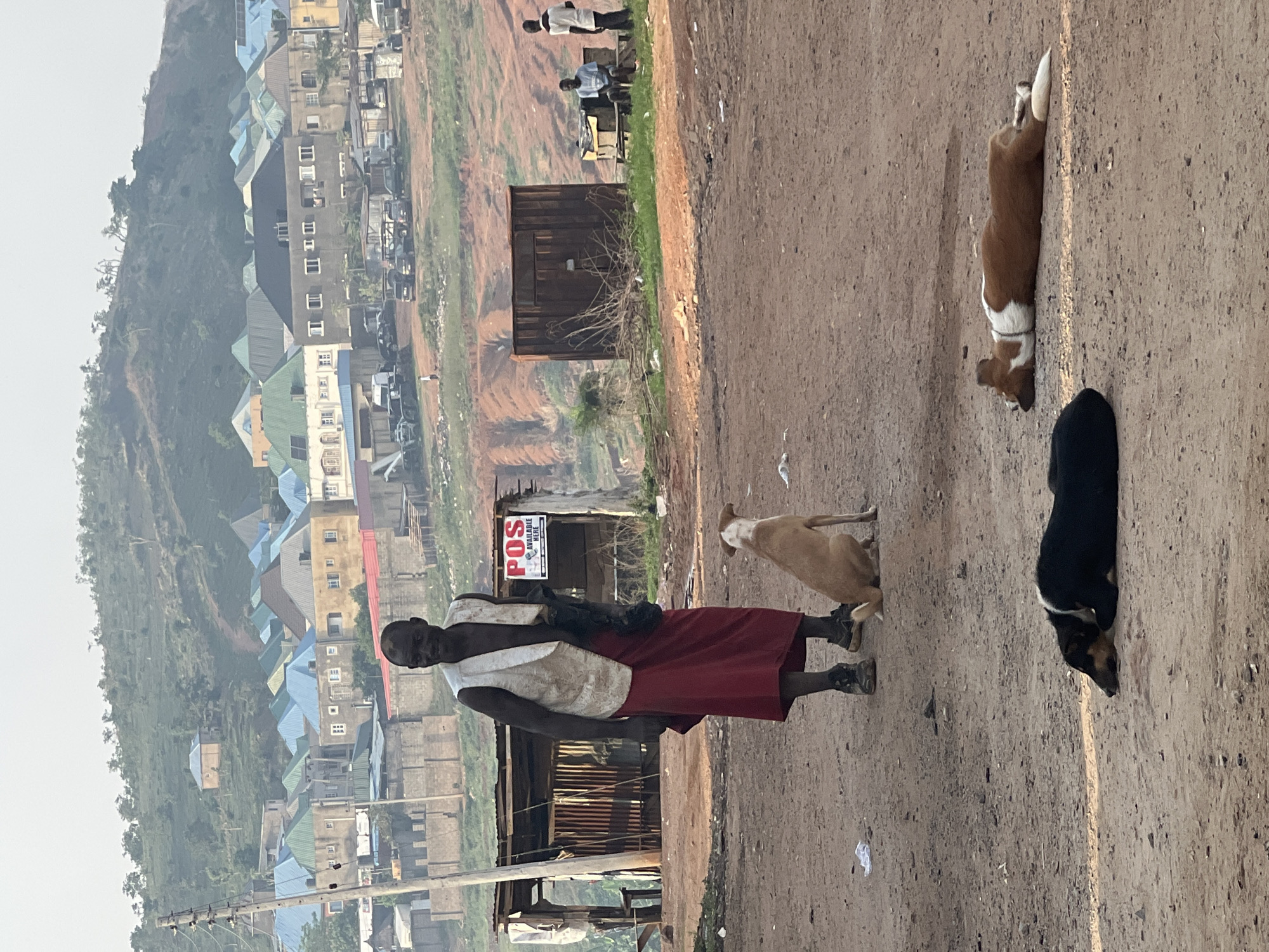 A man in a red skirt and white shirt walks along a dirt path in Abuja, Nigeria. Three dogs are on the path. Behind him are dilapidated wooden structures with a sign reading "POS AVAILABLE HERE" and a view of buildings and a hillside. In the background, two men are standing near a wooden shack.
