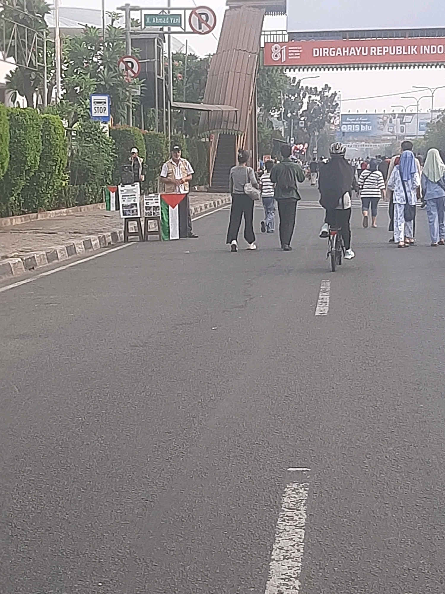 In Bekasi, Indonesia, a street scene unfolds with pedestrians and a bicyclist moving along an asphalt road. On the left side of the road, a vendor stands behind a display of Palestinian flags and informational posters, with a "STOP" sign visible above. In the background, a banner reads "81 DIRGAHAYU REPUBLIK INDONESIA," indicating a national celebration. The environment appears to be an urban setting with trees and buildings in the distance, under an overcast sky.