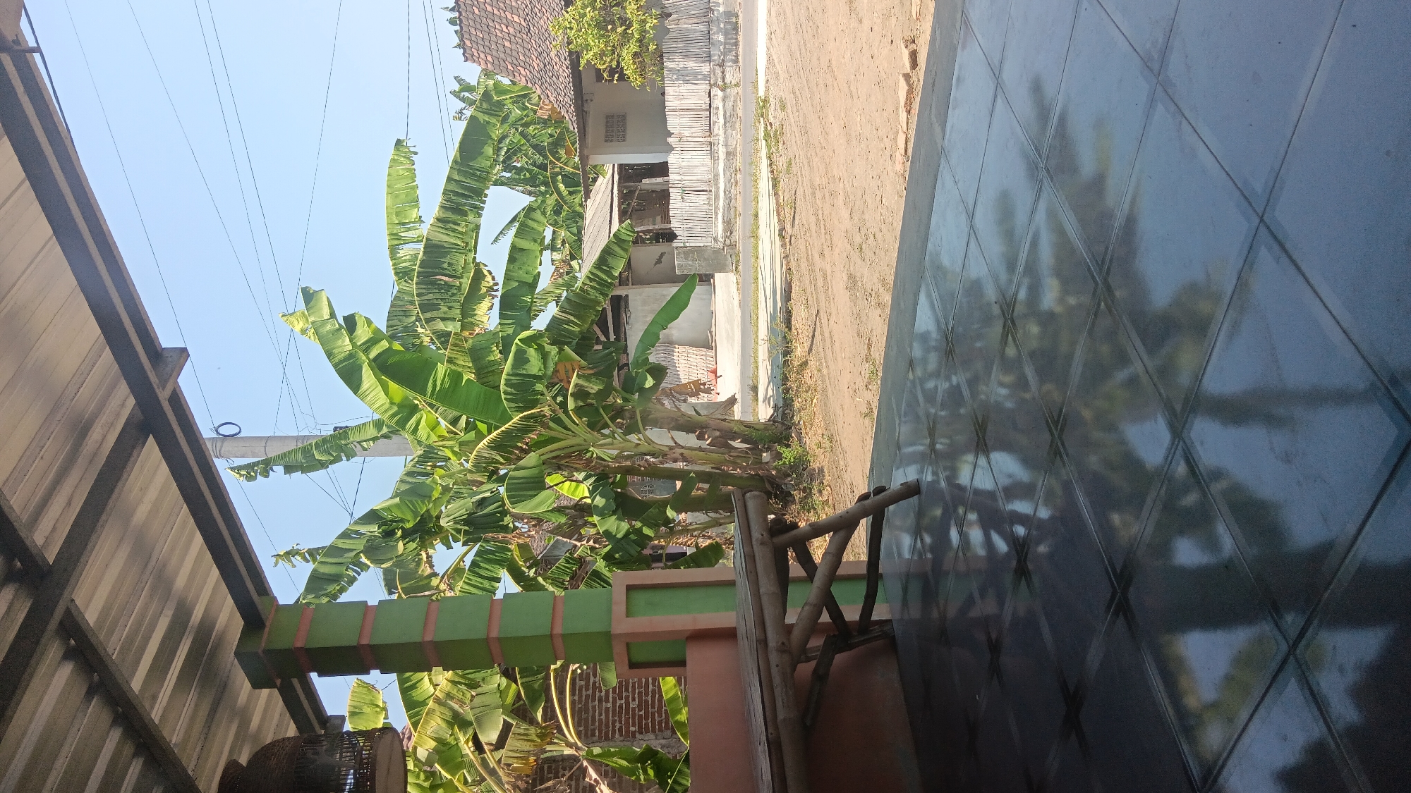 This is a medium-angle, eye-level, outdoor shot taken from under a covered patio area. In the foreground, a tiled floor reflects the surrounding environment. To the left, a bamboo bench sits on the tiled surface, with a supporting pillar visible. The pillar is green with orange accents. Behind the bench, a banana tree with large green leaves is prominent. In the background, there are several houses with tiled roofs and wooden fences. A dirt road and some vegetation are also visible. The sky is clear and blue. The scene depicts a rural or semi-rural setting in Sonobekel, Indonesia. There are no people visible in the image.