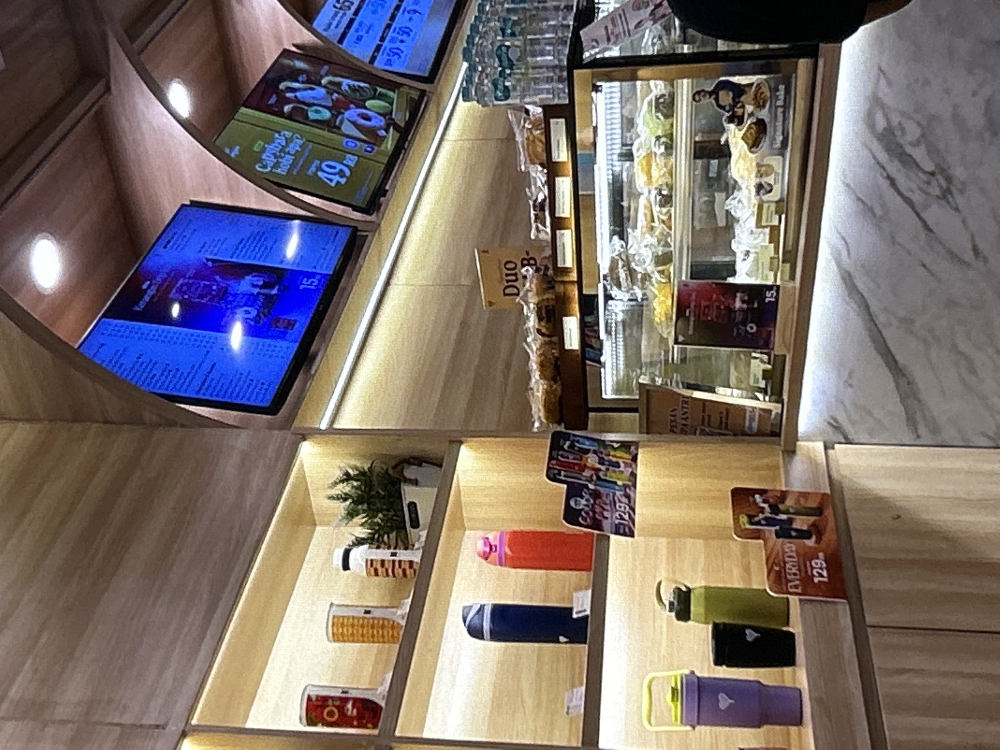 The image shows a display shelf with various items, including bottles, cups, and a small plant. Above the shelves, there are digital screens displaying menus and prices. A glass display case filled with baked goods is visible in the foreground, with a marble countertop in front of it. A person's arm and shoulder are visible on the far right side of the frame, indicating their presence in the scene. The overall environment appears to be a commercial establishment, likely a cafe or a store. The location is identified as Depok, Indonesia.