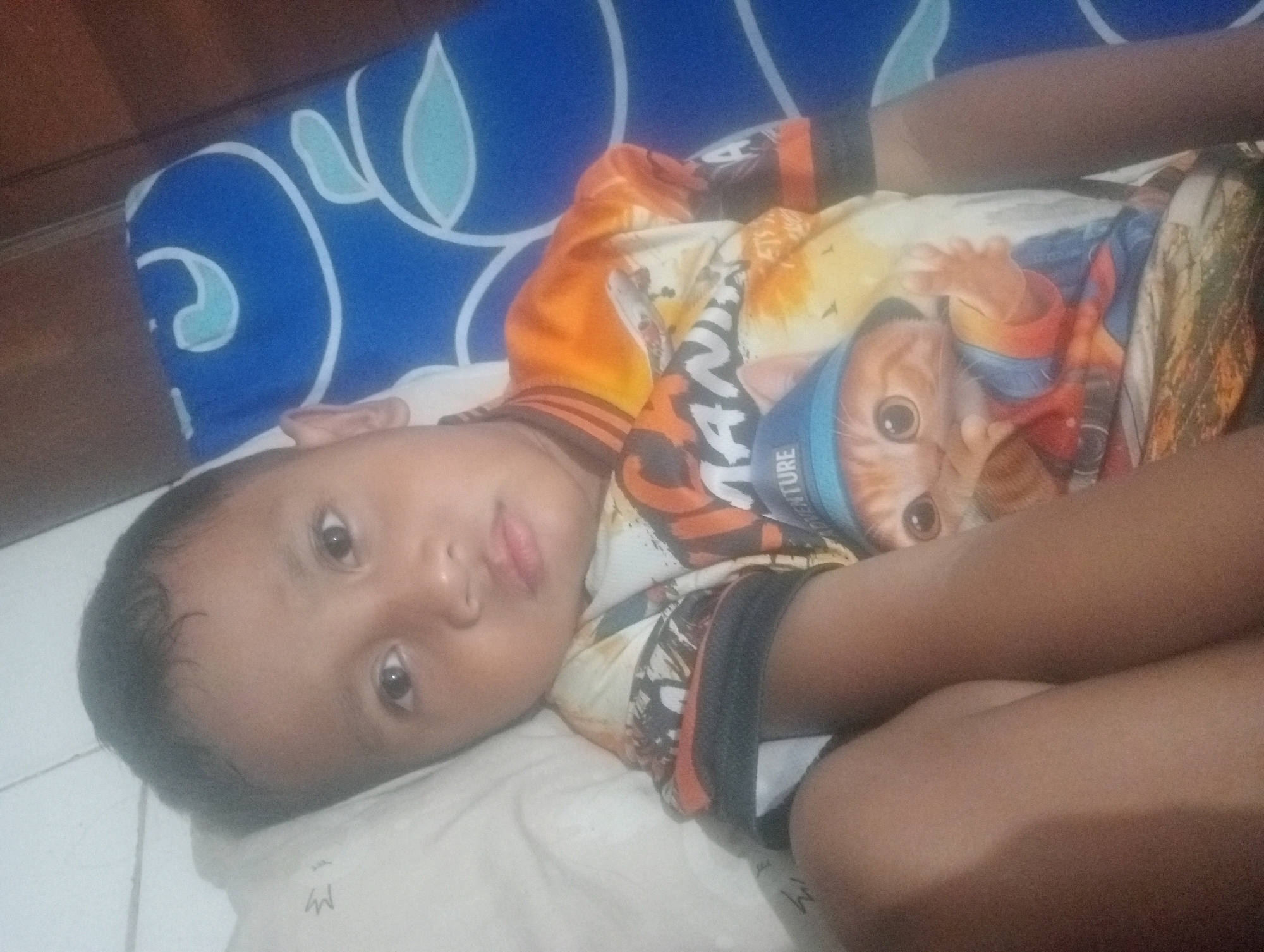 A young child is lying on their side on a white, patterned sheet. The child is wearing a t-shirt with a graphic of a cat on it. The child's face is visible, and they appear to be looking directly at the camera. The background consists of a blue and white patterned pillow. The setting appears to be indoors, possibly on a bed or couch, with wooden floor visible to the left. The image is taken from a high angle.