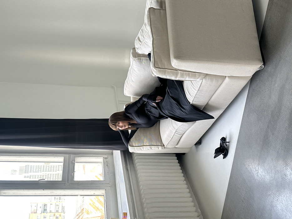 An adult female, with chin-length brown hair and bangs, reclines on a light beige fabric sofa in an indoor setting. She is dressed in a black long-sleeved top and a flowing black lower garment, looking contemplatively towards a large multi-paned window on the left side of the frame. Her left hand rests near her head, while her right hand is obscured by fabric near her lap. On the floor in front of the sofa, a pair of black high-heeled shoes lies discarded.
The room features white walls, a white horizontal radiator beneath the window, and a dark curtain bordering the window's right side. The floor is bifurcated by a diagonal line, presenting a white section near the window and a light gray section beneath the sofa and extending into the foreground. Outside the window, a generic urban landscape of multi-story buildings, including a taller structure with numerous windows, is visible under an overcast sky. While the image indicates an urban environment in Moscow, Russia, specific landmarks allowing precise location identification are not visible.