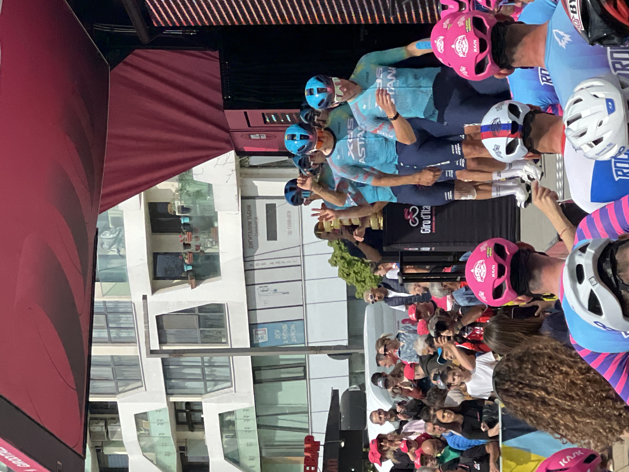 The image depicts a group of cyclists, potentially a team, posing for a photograph. Most of the subjects wear cycling gear, including helmets of varying colors (pink, white, blue) and matching teal cycling jerseys. They are standing on a platform near a crowd of spectators, who are taking photos and observing.
In the background, buildings can be seen, indicating an urban setting. A sign in Bulgarian reveals that the scene is taking place in Burgas, Bulgaria, specifically near a "Center for X-ray diagnostics". A sign with the Giro d'Italia logo is also visible. The sky is not visible in the image.