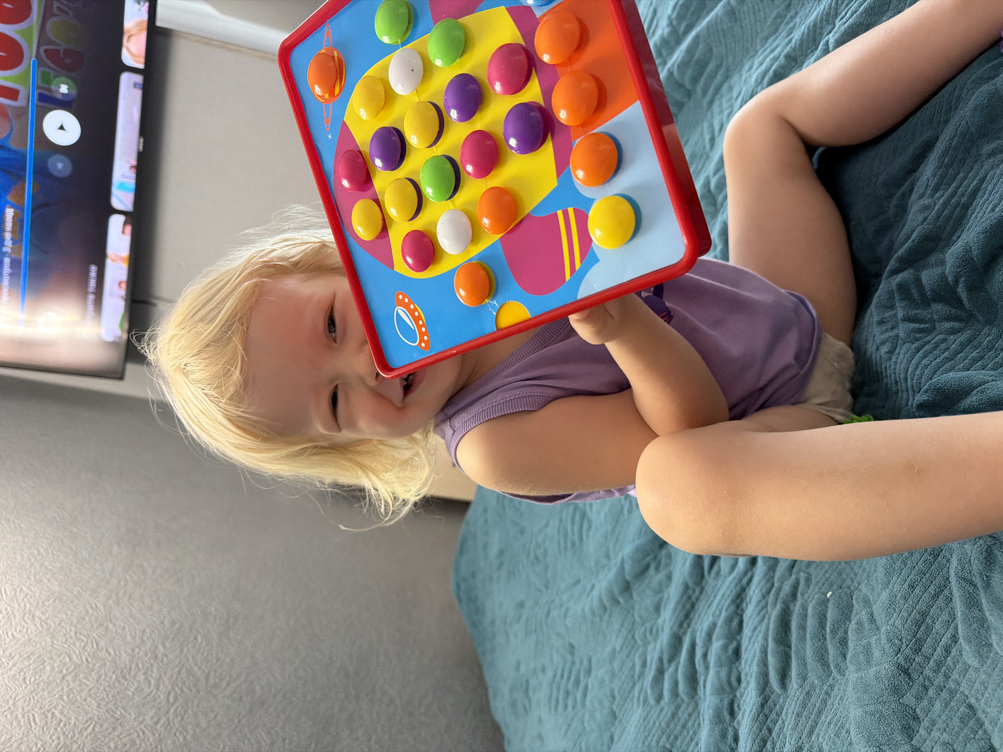 A young child with blond hair is lying on a bed with a teal, textured blanket. She is holding a colorful toy with round buttons. The child wears a purple shirt and light-colored shorts. A TV is visible on the left side of the image, displaying a menu with various icons and text in a language possibly Russian. The setting is indoors with a gray wall visible in the background. There is no information given that would connect the image to the city of Sochi, Russia.