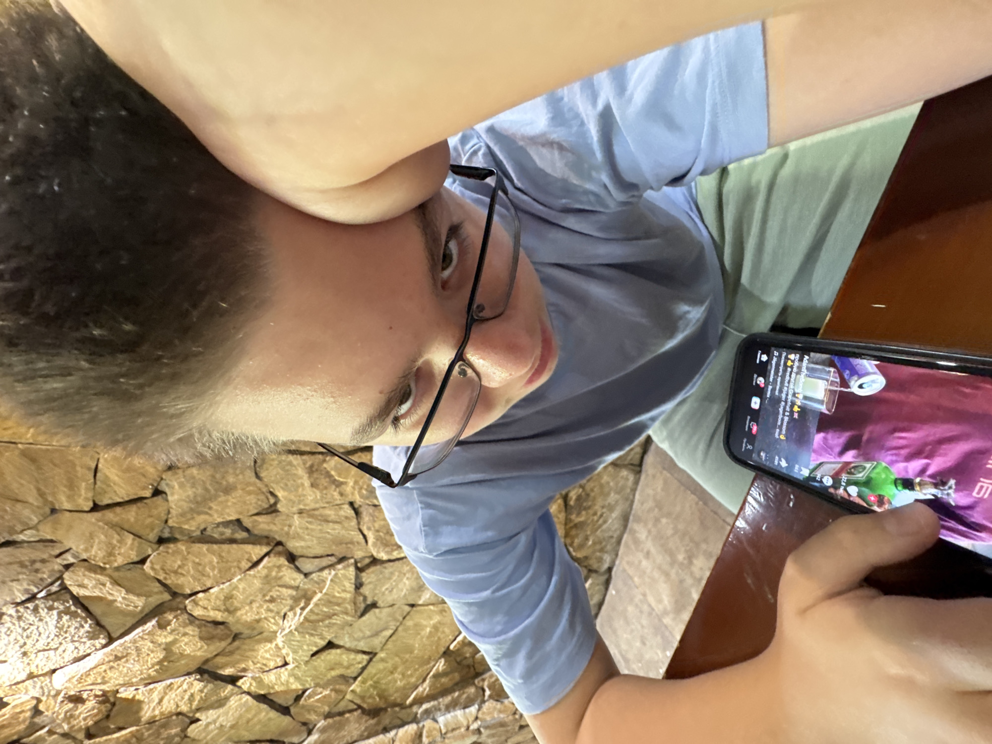 A young boy with short, spiky brown hair and glasses is leaning his head on his right arm, looking down at a smartphone in his left hand. He is wearing a light blue t-shirt and light green shorts. The background consists of a textured stone wall. The phone screen shows a video with a green bottle and a purple background. The boy is indoors, likely at a table, suggested by the brown surface in the foreground. The image does not contain information about the location in the city of Sredets, Bulgaria.