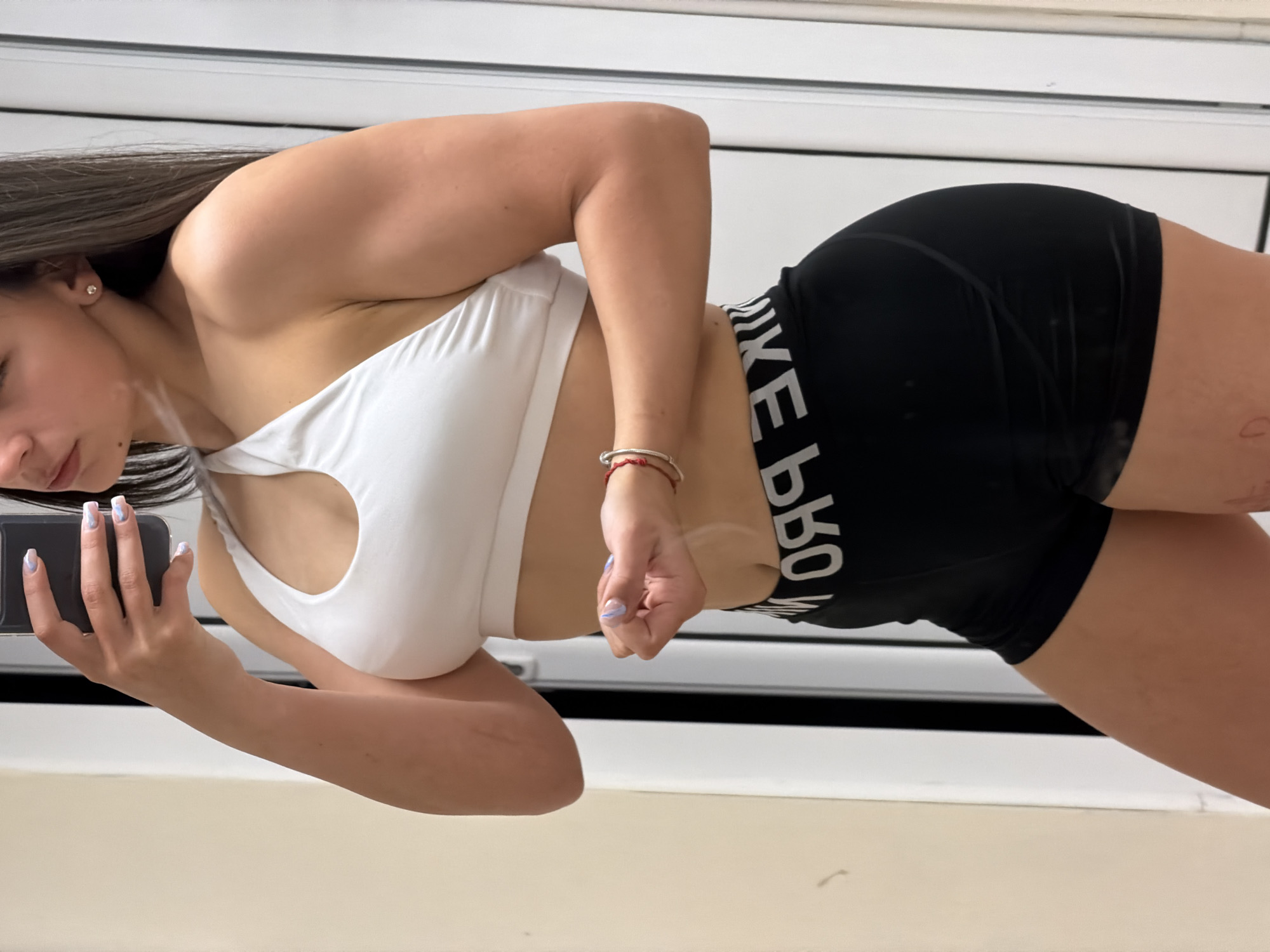 A woman is taking a selfie in a room with white walls and a window with horizontal blinds. She is wearing a white sports bra and black athletic shorts with the words "NIKE PRO" printed on the waistband. She is holding a smartphone in her left hand, and her right hand is near her waist. She has long, dark hair. The location is not identifiable as Gabrovo, Bulgaria.